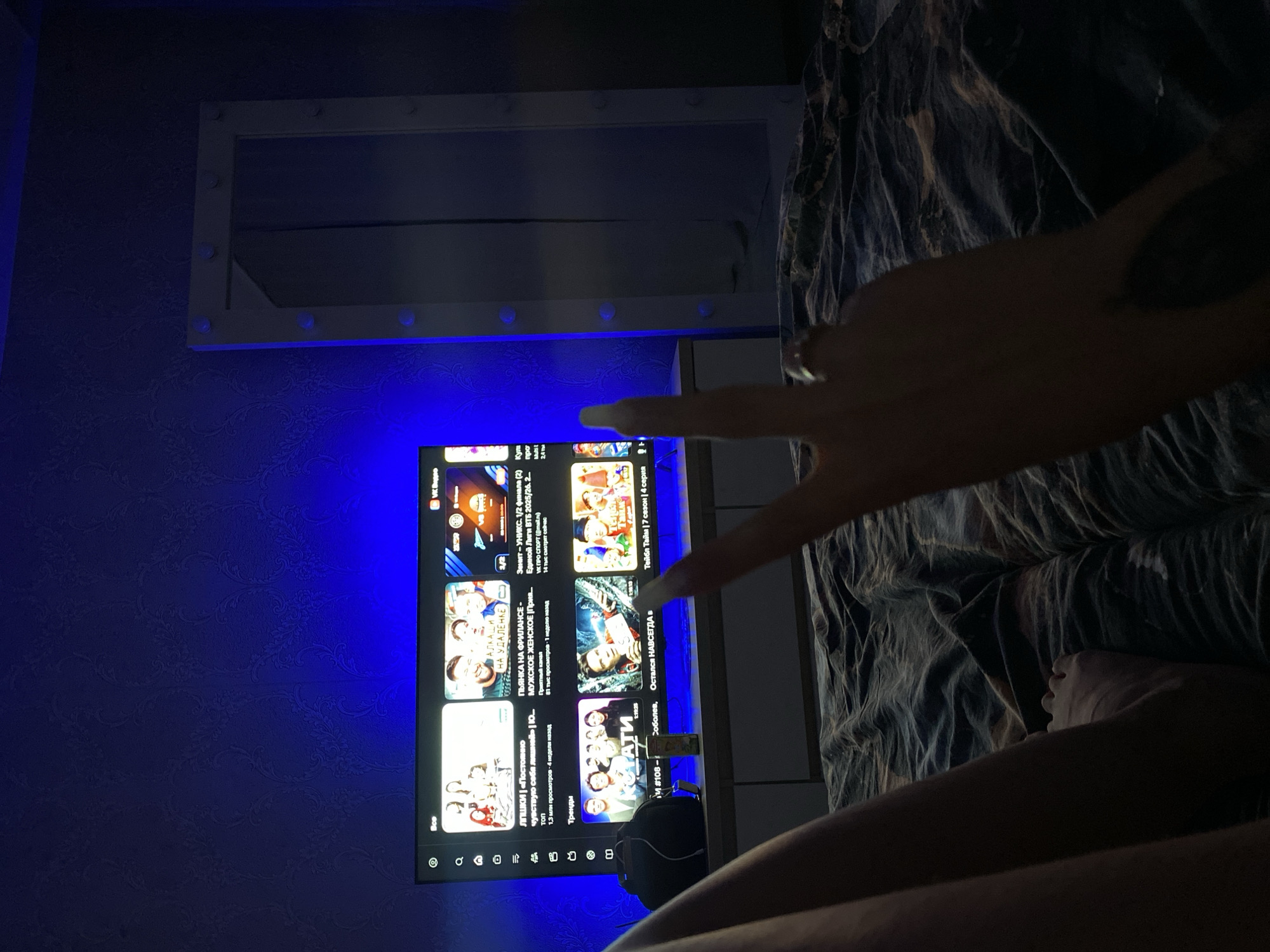 A person is lying in bed in a dimly lit room with blue ambient lighting, possibly in Chaikovsky, Russia. A hand is reaching towards a television screen displaying a YouTube interface with Russian text. Above the television, there's a mirror with vanity lights. The person's legs and feet are visible in the foreground on a dark patterned bedspread. There is a black bag next to the television.