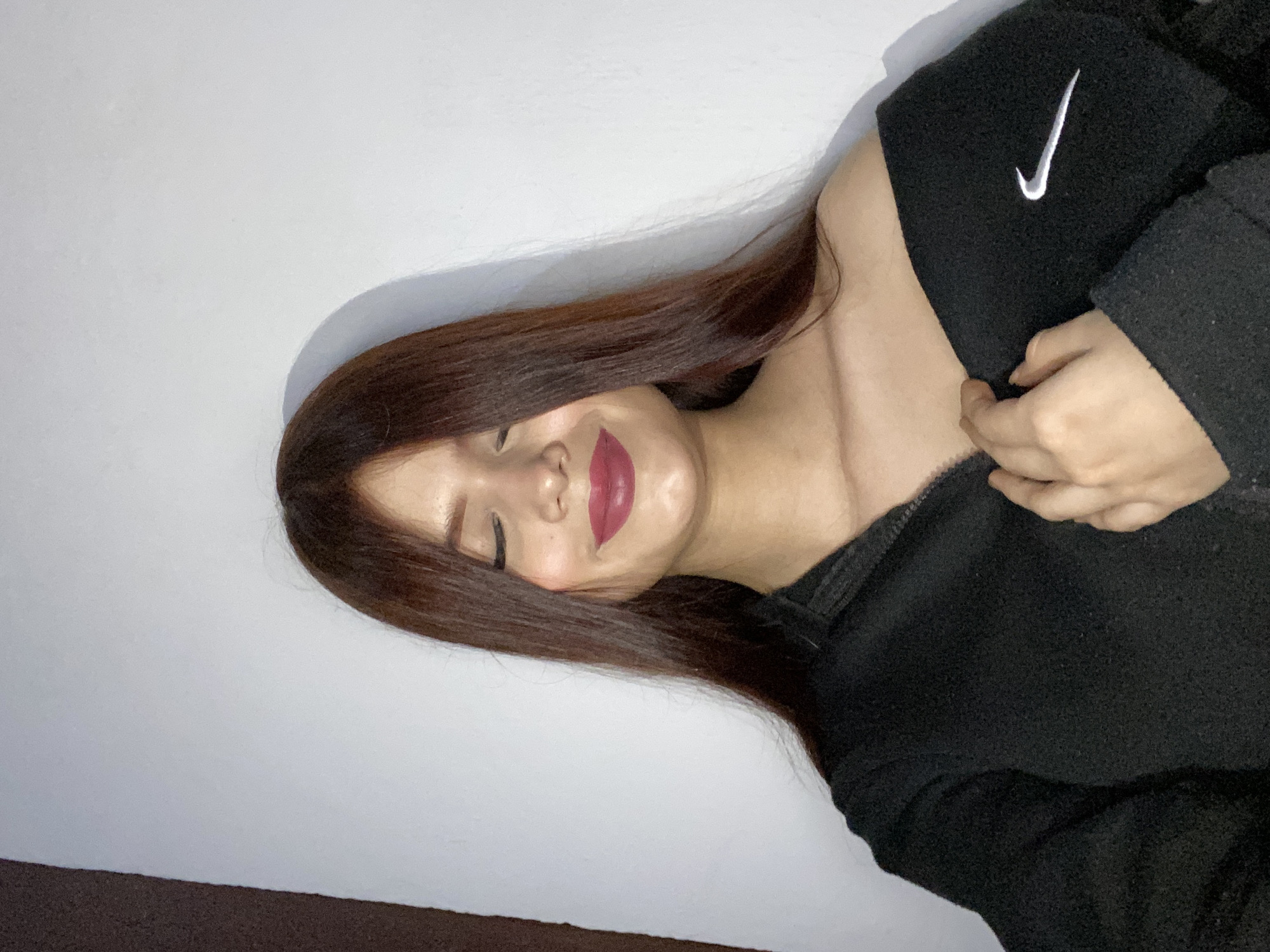 A woman with brown hair is lying down, wearing a black jacket with a white Nike logo. She has her eyes closed and is wearing red lipstick. The background is a plain white wall. The location is unknown and there is no evidence to support the presence of Juan Griego, Venezuela.