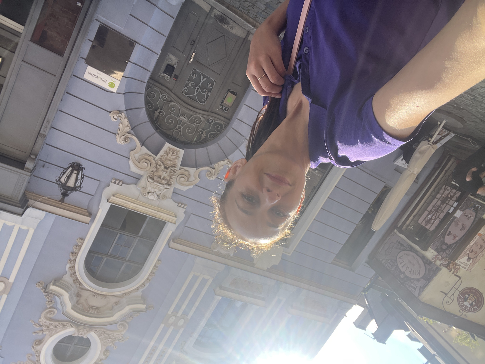The image shows a woman taking a selfie in front of a building with ornate architecture. She is wearing a purple polo shirt and has a pink strap from a bag visible over her shoulder. Her hair is pulled back, and she is smiling. The building behind her is painted a light blue and features intricate decorative elements around a arched doorway and windows. A street lamp is mounted on the building. Signs are visible on the building, one of which appears to be for a coworking space. The ground is paved with cobblestones. The sun is shining brightly, creating lens flare in the upper part of the image.