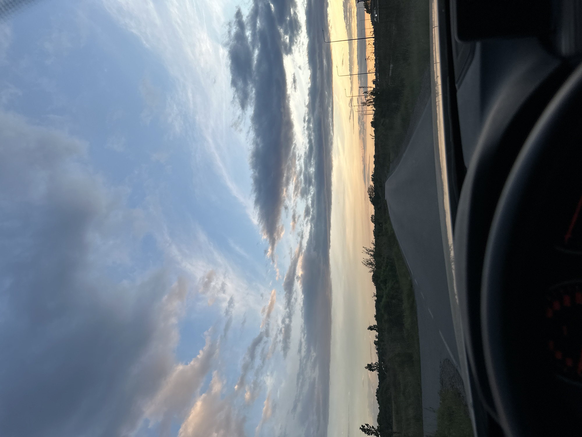 The image is taken from inside a vehicle, likely a car, with the steering wheel partially visible on the right side. The view is of a road stretching into the distance on the right, with trees and vegetation lining its side. Beyond the road and trees, the sky dominates the scene. The sky displays a gradient of colors, with hues of blue, grey, orange, and yellow, likely indicating either sunrise or sunset. Clouds are present, some dark and dense while others are lighter and more diffused. There are streetlights in the distance.
Unknown, Unknown is not discernible from the image.