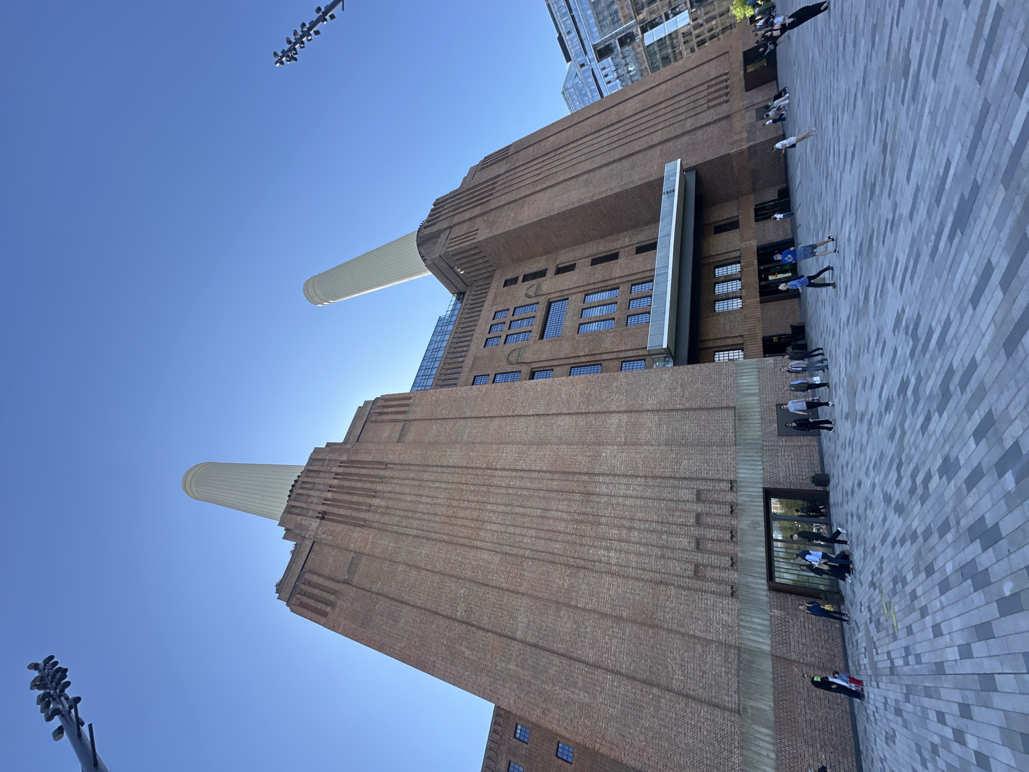 The image shows a low-angle view of Battersea Power Station in London, United Kingdom, under a clear blue sky. The brick facade of the building is prominent, with two of its iconic chimneys visible. People are walking on a paved area in front of the building. A modern building is visible in the background, to the right of the power station. A sculptural light fixture is in the lower left corner.