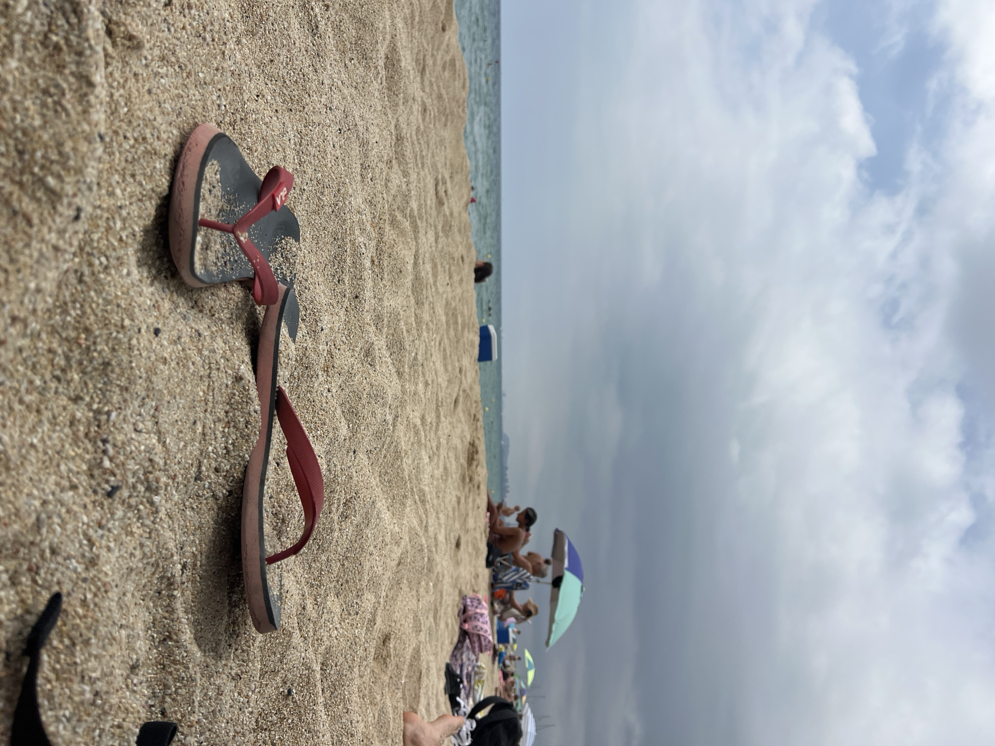 The image is a low-angle, close-up shot of a pair of red and black flip-flops lying in the sand on a beach. The sand is the primary focus of the foreground. In the mid-ground, the ocean is visible with gentle waves. A few people are scattered along the shoreline, some under umbrellas, enjoying the beach. In the background, the sky is cloudy with a mix of light and dark gray clouds. The overall scene suggests a casual day at the beach, likely in a populated area given the presence of multiple beachgoers and umbrellas. The location is identified as the city of El Masnou, Spain.