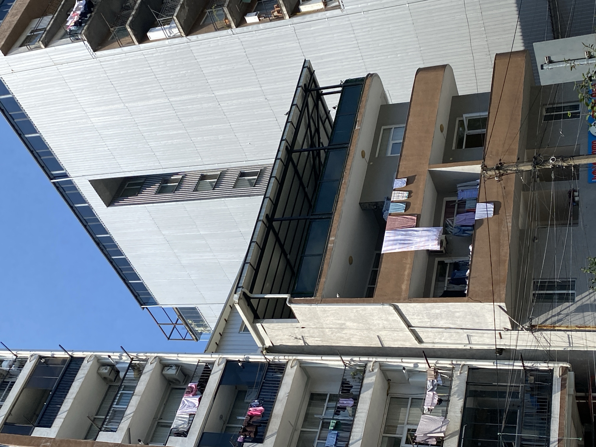 This image depicts a street scene in Batumi, Georgia, featuring a cluster of apartment buildings under a clear blue sky. The buildings are varied in their architecture and construction. On the left, a light-colored, multi-story apartment building displays laundry hanging from balconies on multiple levels. Air conditioning units are visible on some balconies. Adjacent to it, another building with a brown facade also shows laundry draped over railings, with a covered balcony area above. To the right, a tall, modern building with a corrugated metal exterior dominates the background. Numerous wires and utility poles are visible in the foreground, suggesting an urban environment. The scene is devoid of people.