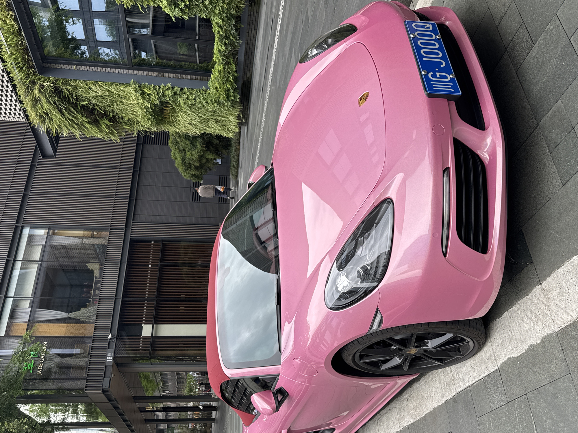 A pink Porsche sports car with a red convertible top is parked on a paved area in what appears to be an urban setting. The car's license plate reads "川G·J000Q." Behind the car is a modern building with a sign that reads "Jade Garden." The building has vertical wood-like slats and green foliage growing on its facade. A person is walking away from the camera in the background, near the foliage. The location is Jinjiang District, China.