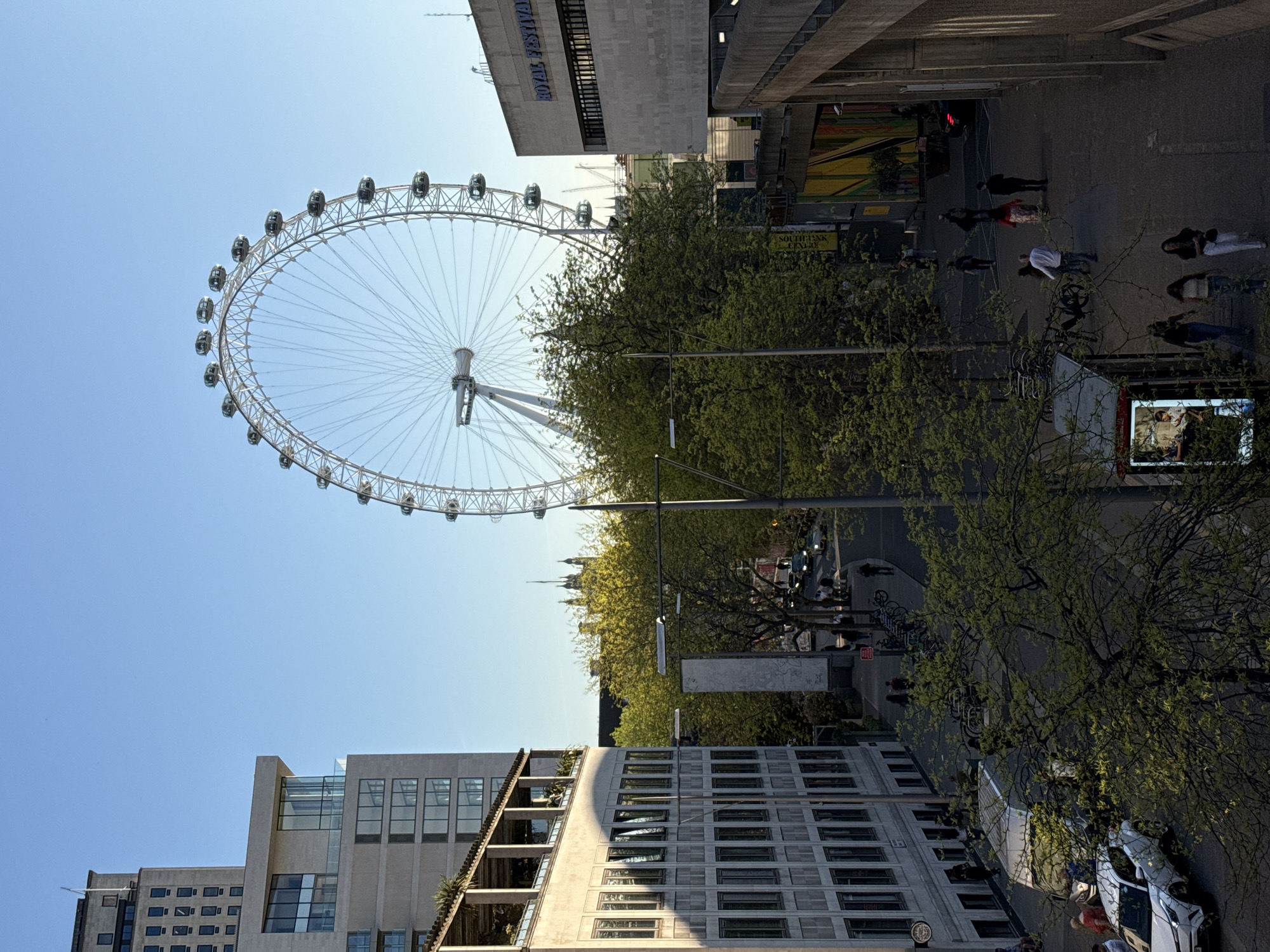 The image depicts a sunny, daytime scene in London, United Kingdom, featuring the London Eye Ferris wheel prominently in the upper left portion. The wheel is surrounded by a clear blue sky. Below the London Eye, trees line a street with pedestrians walking along the pavement. Vehicles are visible on the road. Buildings, including what appears to be the Royal Festival Hall, frame the sides of the image. People are walking, some are in groups. The overall environment suggests a bustling urban landscape with iconic London landmarks.