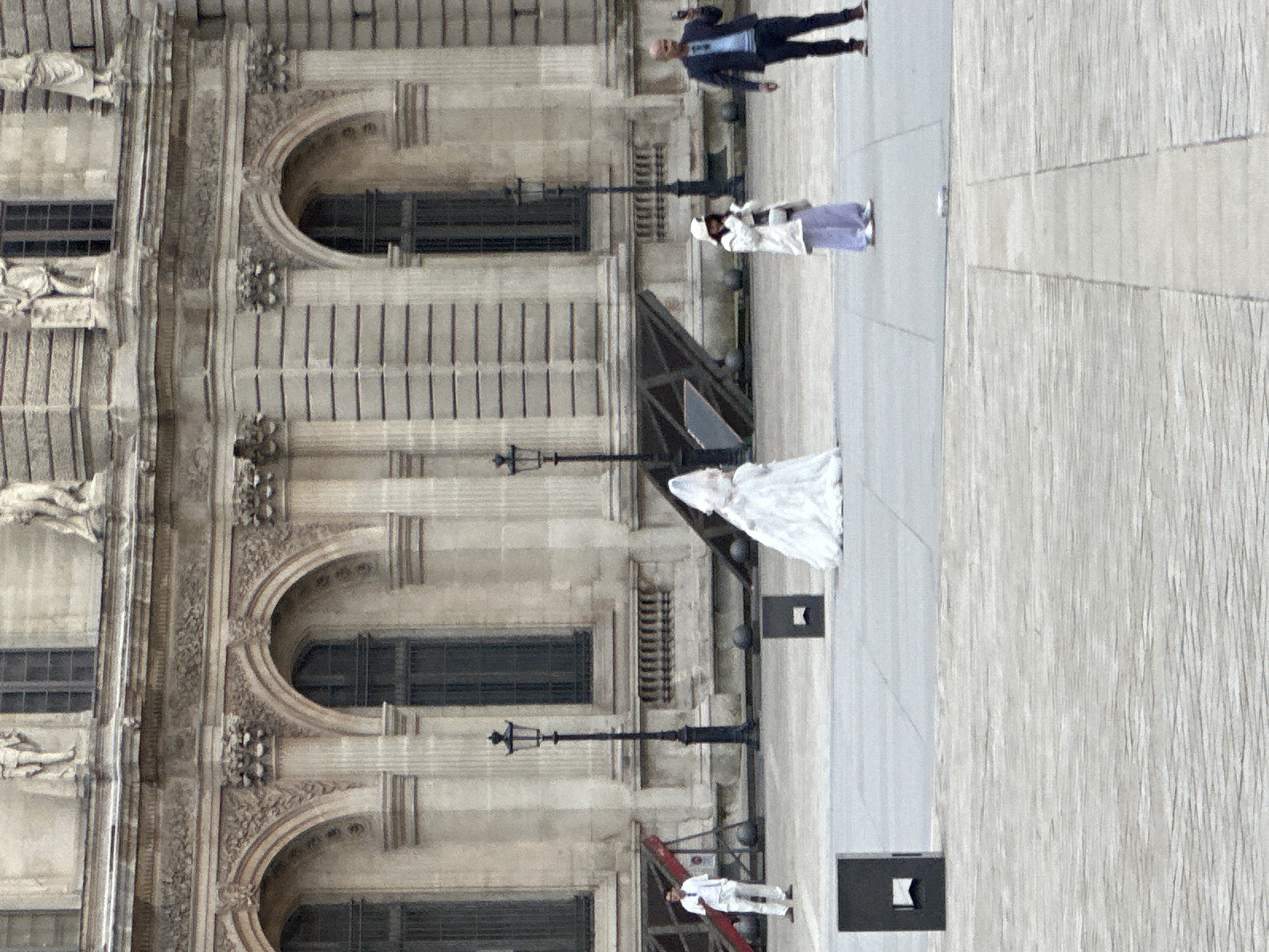 The image shows the Louvre Museum in Paris, France. The museum's exterior is visible, with its distinct arched windows and ornate architectural details. Several people are present in the scene. A person in a white dress stands near a temporary structure. Another person in white clothing and a hat stands by a ladder. Further up, a person in a light-colored coat and purple skirt is seen, and a man in a dark suit walks nearby. The ground is paved with light-colored stone. Streetlights line the area in front of the museum.