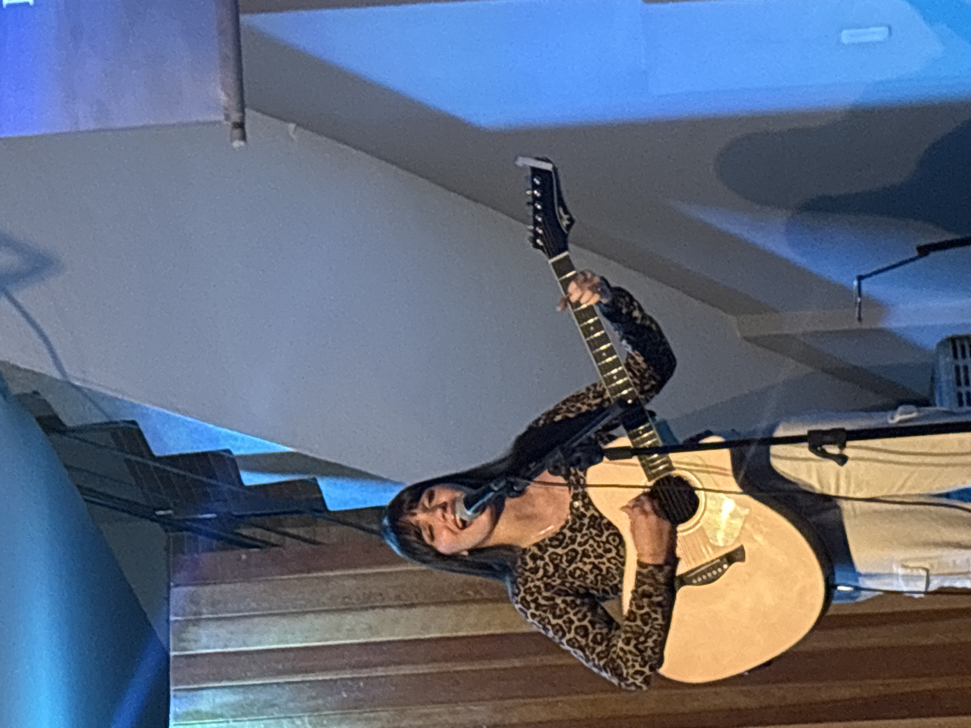 An image shows a woman singing and playing an acoustic guitar in a room. The woman has long dark hair and is wearing a leopard-print shirt and white pants. She is standing in front of a microphone and a light blue wall. The room has wooden walls on the left side and a slanted white wall on the right side. There is a shadow of a person projected on the blue wall. The overall lighting appears to be from stage lights, with a strong blue hue on the right side of the image. The scene is consistent with a live performance or practice space, and there is no clear indication of the specific location within São Paulo, Brazil.