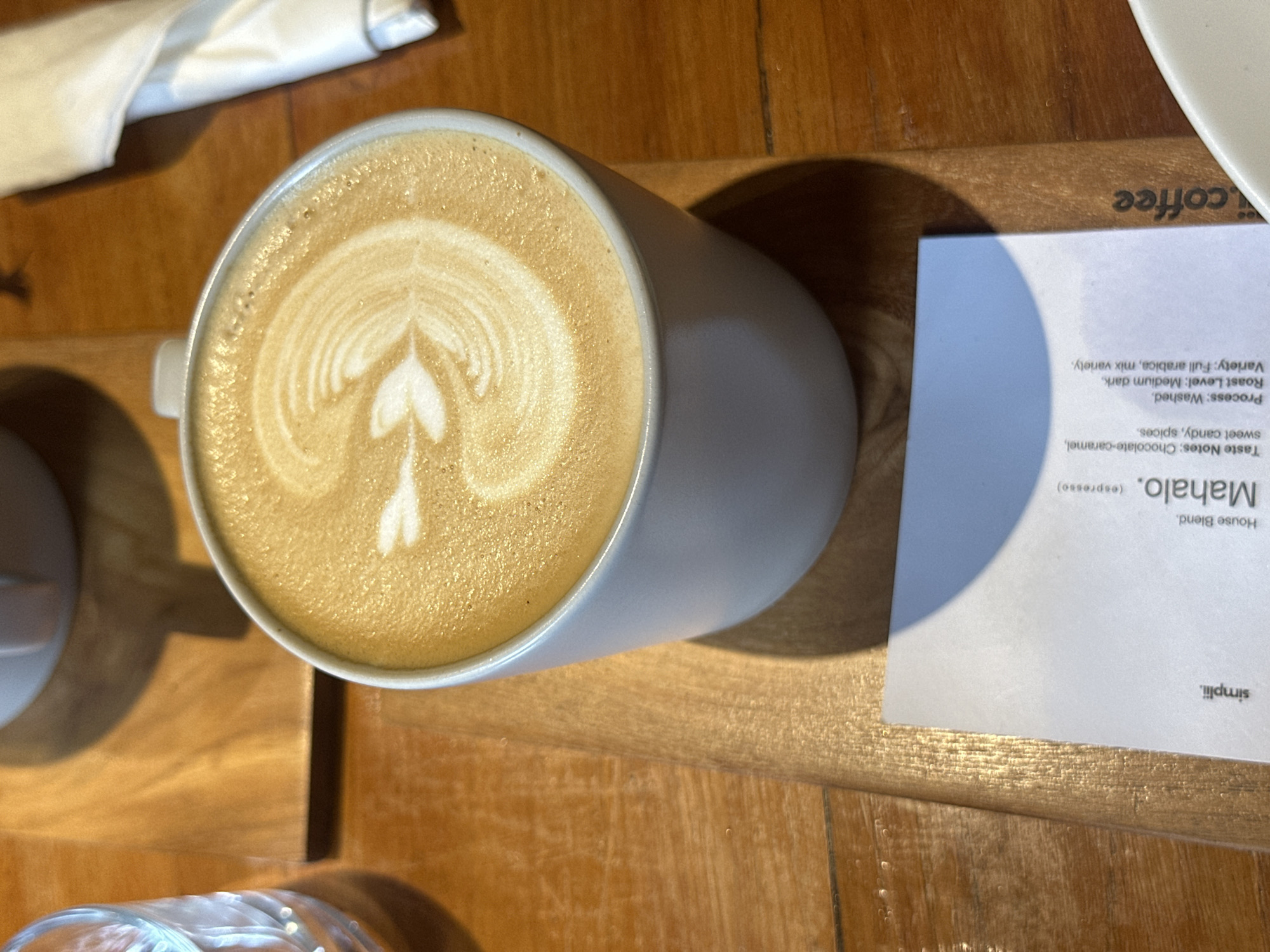 A high-angle, close-up shot shows a cup of latte art coffee. The coffee is in a white mug with foam art resembling a leaf or feather. The mug sits on a wooden tray. To the left of the mug is a clear glass with a ribbed design, containing a light-colored liquid. To the right, a white napkin is folded. In the foreground, a white card displays text related to the coffee, including "simplii.", "Mahalo. (espresso)", "House Blend", "Taste Notes: Chocolate-caramel, sweet candy, spices.", "Process: Washed", "Roast Level: Medium dark", and "Variety: Full arabica, mix variety.". The background is a wooden surface.