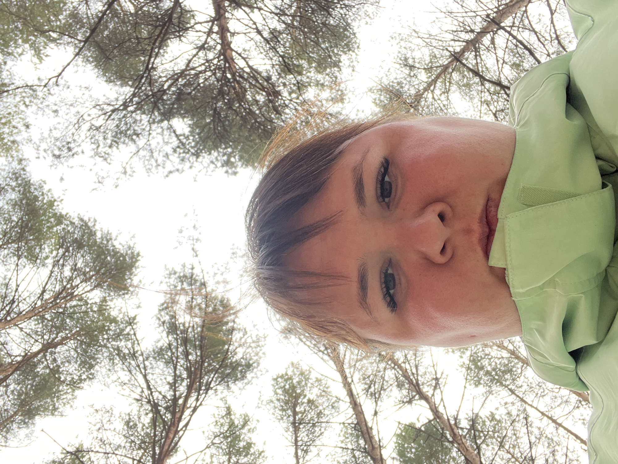 The image presents a unique low-angle self-portrait or selfie, looking upwards. The main subject is a person, likely female, whose face and upper chest are prominently featured in the lower right portion of the frame. Her face is turned, and she looks directly into the camera with a neutral to slightly pensive expression. She has light brown hair and is wearing a light green jacket, with a velcro fastening visible on the collar area.
The background consists of a dense canopy of tall trees, characteristic of pine or similar coniferous species, with their branches and sparse foliage silhouetted against a bright, overcast sky. This upward perspective suggests the photo was taken either while the subject was looking up from the ground or holding the camera above them, pointing down towards themselves while capturing the sky. The lighting is bright and diffused, indicating an overcast day. The scene depicts an outdoor environment, specifically a forest or wooded area in Chaikovsky, Russia. There is no visible text in the image.
