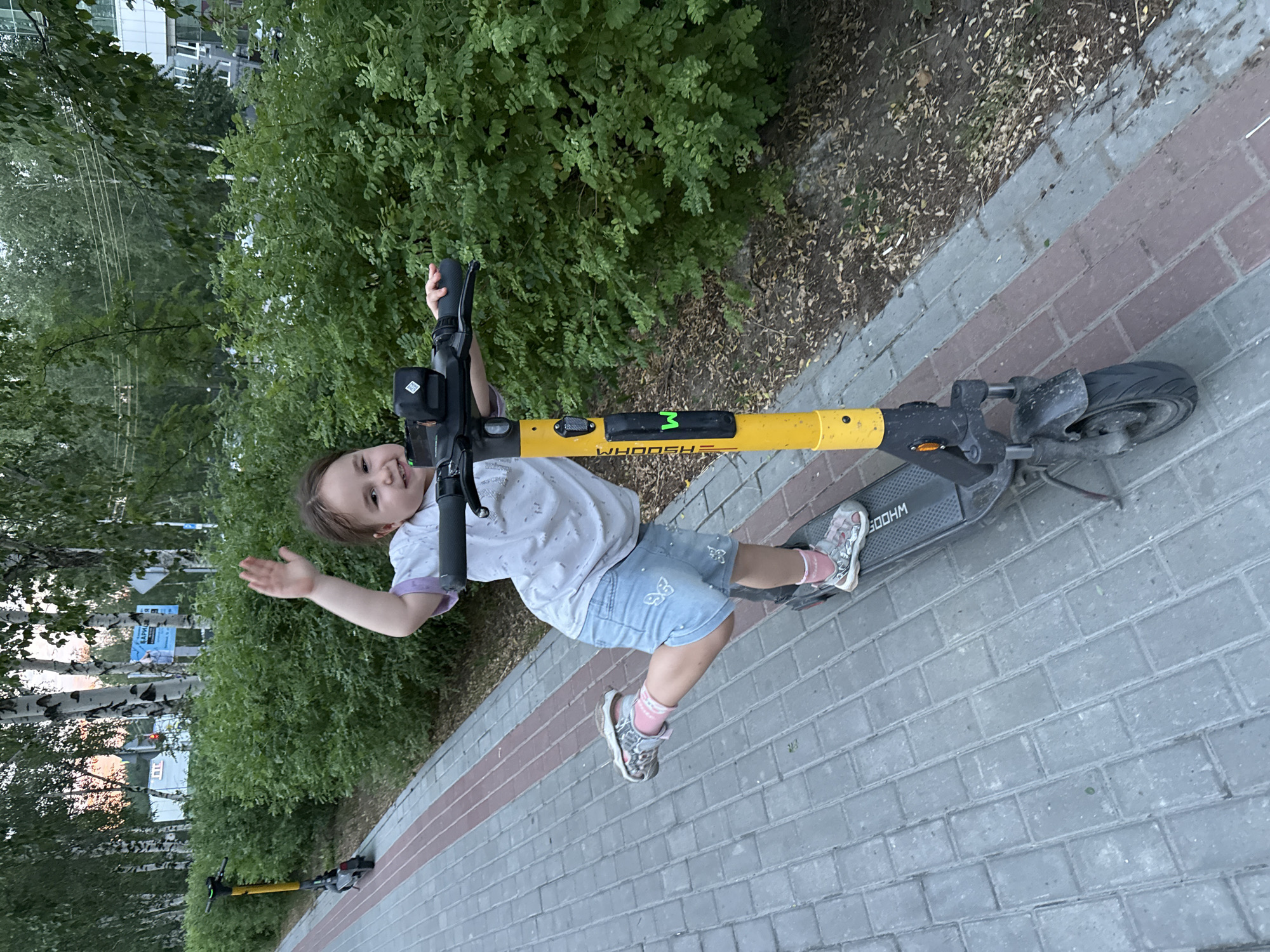 A young child with dark hair, wearing a white t-shirt with purple trim, blue denim shorts adorned with butterflies, pink socks, and grey sandals, is playfully posing on a yellow and black electric scooter. The scooter has the brand name "WHOOSH" printed on its handlebars. The child is holding onto the handlebars and waving with their left hand, looking towards the camera with a cheerful expression. The scene is outdoors, with a paved walkway made of grey and red brick-like tiles. To the left of the walkway is a lush green bush, and behind that, there are birch trees with white trunks and green foliage. In the background, glimpses of buildings and what appear to be posters on poles can be seen. The overall impression is a bright, daytime scene, likely during pleasant weather, given the child's attire. The location context is Surgut, Russia.