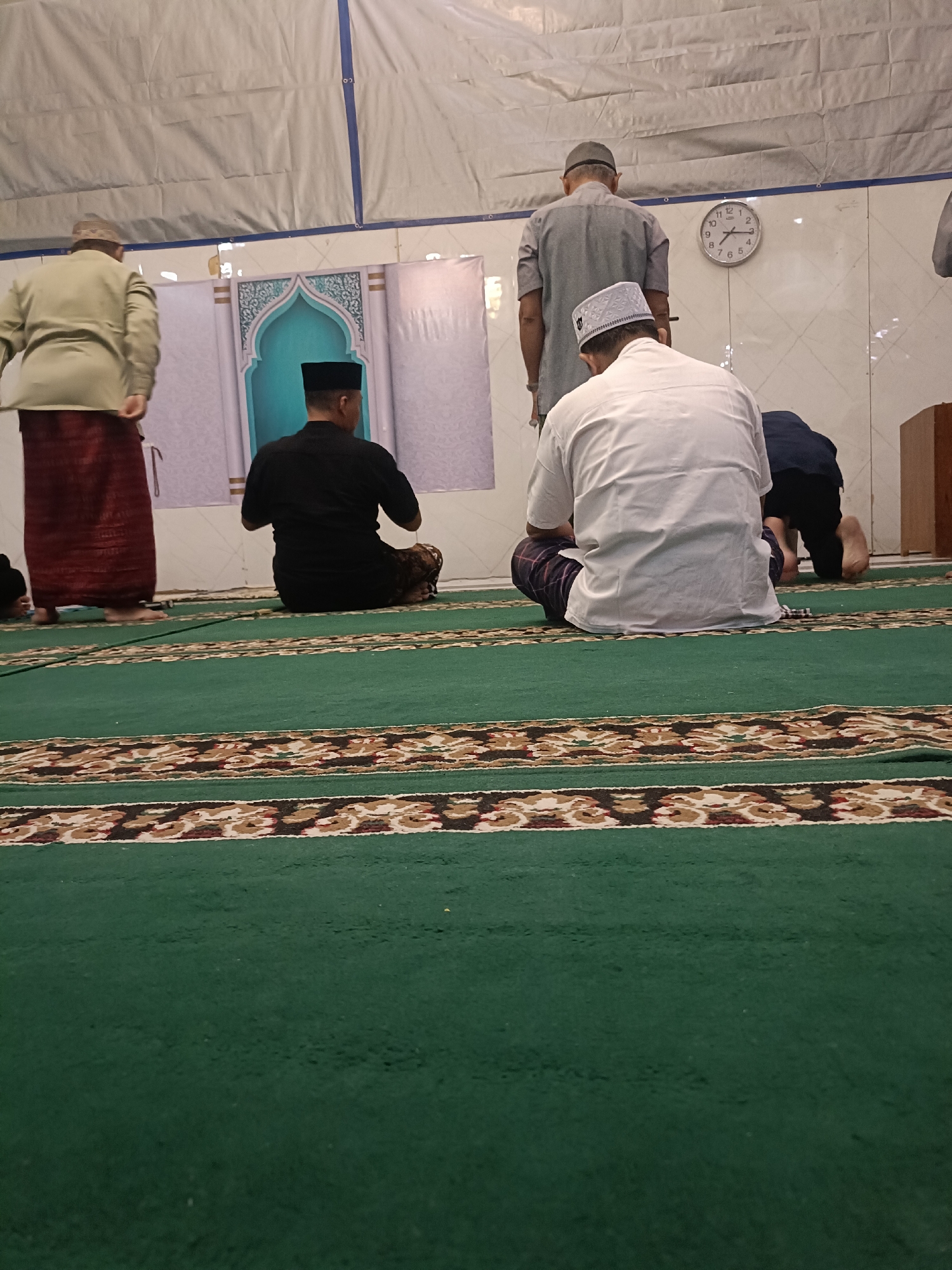 The image depicts a group of Muslim men praying inside a mosque. The prayer hall features a green carpet with decorative borders, and a wall with an arched motif. A clock on the wall indicates it is around 9:15. Several men are in various stages of prayer, facing the qibla. One man in the foreground is wearing a white shirt and a white cap, seated on the carpet. To his left, another man in a black shirt and cap is also seated. Further back, a man in a grey shirt and cap stands, and another man in a light-colored shirt and patterned skirt is on the far left. The lighting suggests it is evening or indoors. This scene is consistent with the interior of a mosque in Kediri City, Indonesia, where Islamic prayer is a common practice.