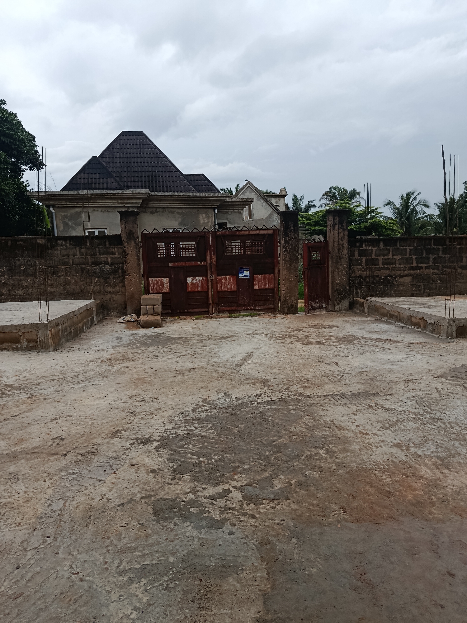 The image depicts an overcast day in Ukpor, Nigeria. The foreground is a concrete courtyard with some patches of dirt and debris. To the left, there is a partially constructed concrete foundation with rebar protruding. A dark brick wall runs horizontally across the midground, separating the courtyard from the property behind it. Centered in the image is a large, rusty, double-leaf metal gate, with a smaller, single-leaf gate to its right, slightly ajar. The gate is set between two large, weathered concrete pillars. Behind the wall and gate, a house with a distinctive dark, multi-faceted roof is visible, along with other structures and lush green trees, including palm trees. The sky is heavily clouded.
