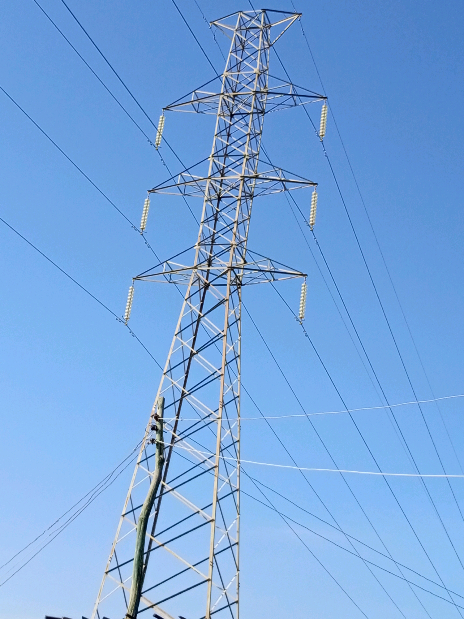 This is a vertical, full-frame, medium shot of a tall, metal electricity pylon against a clear, bright blue sky. The pylon is made of a lattice of metal beams and supports multiple power lines. Three sets of insulators, each with multiple stacked, translucent discs, are visible hanging from the crossbars of the pylon, holding the power lines. A thick, green-covered cable is attached to a wooden pole that is leaning precariously against the metal pylon's structure. The wooden pole is positioned to the left of the main pylon, and it appears to be supporting some of the power lines. The scene is set on Rukuba Road in Nigeria, indicating an outdoor, possibly urban or semi-urban environment. The image has a slightly grainy texture.