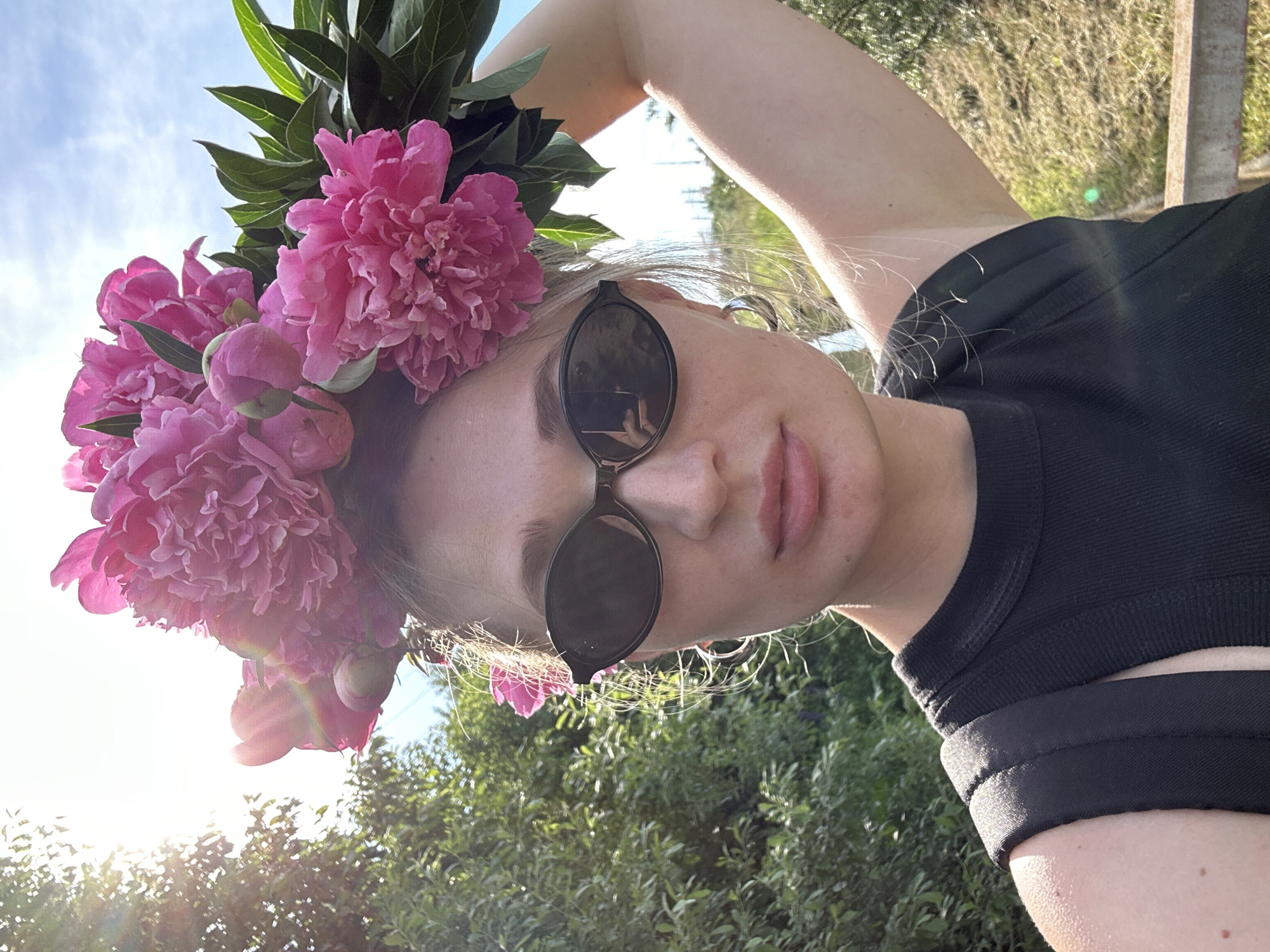A low-angle, close-up, vertical shot captures a young woman wearing a crown of pink peonies and black sunglasses. She is outdoors, with a bright sky and greenery visible in the background, suggesting it is daytime and likely warm weather. The woman has light-colored hair and is wearing a black, ribbed top with cutouts on the shoulder. Her right arm is raised and bent behind her head. The sun is shining brightly, creating lens flare in some areas of the image. The peonies are large and vibrant, with some buds and green leaves. The overall mood is relaxed and summery. The location is likely a garden or park in Chaikovsky, Russia, given the sunlight and foliage.