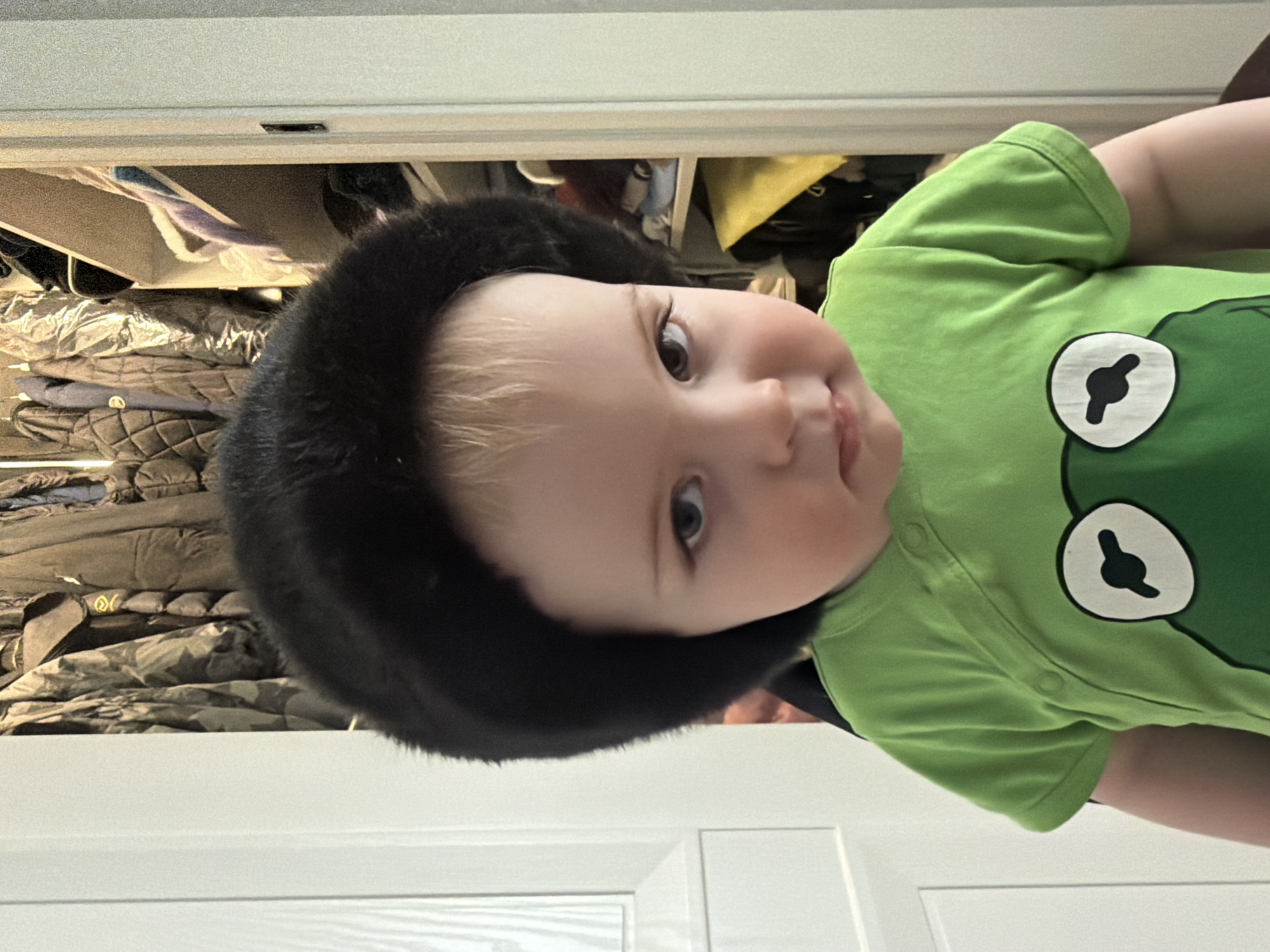 A baby wearing a black fur hat and a green frog-themed onesie is lying down. The background consists of a closet with clothes inside and a white door. The location is not apparent.