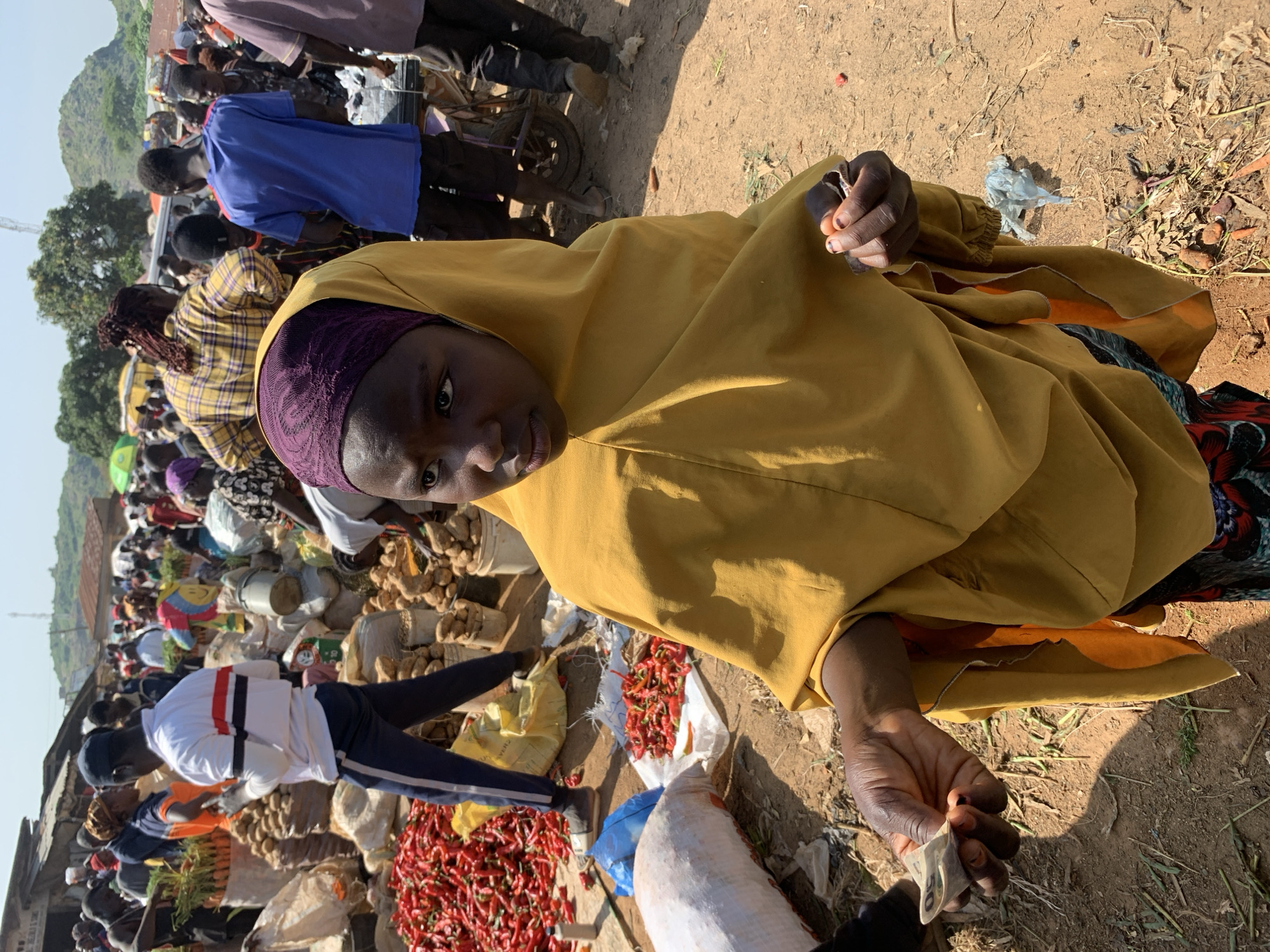 The image depicts a bustling outdoor market scene in Gura Top, Nigeria. In the foreground, a young girl wearing a yellow headscarf and dress is partially visible, her gaze directed to her right. She holds some currency in her right hand, with her left hand outstretched. The background is filled with a variety of market stalls and people. To the left of the girl, large piles of red chili peppers are visible, as well as sacks of produce. In the center background, people are milling about, some near a motorcycle and others near piles of root vegetables. The ground is dirt, and the sky is a bright, clear blue. The overall atmosphere suggests a vibrant and active marketplace.