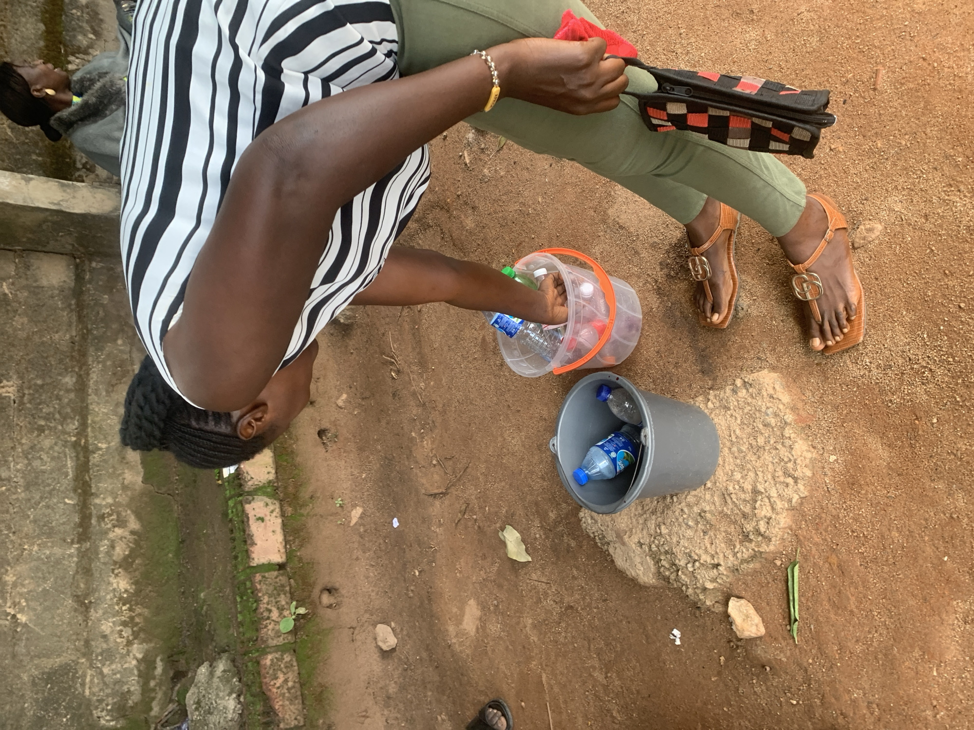 The image shows an overhead view of a person wearing a black and white striped shirt and olive green pants, sitting on the ground. They are holding a clear plastic bucket filled with water bottles and a red cloth. Next to them is a grey bucket with a few water bottles inside. The ground appears to be made of dirt and sand, with some patches of greenery and scattered rocks. The scene is outdoors, with natural lighting suggesting it is daytime. The person's focus is on the contents of the bucket. The overall setting appears to be a casual outdoor environment, possibly a residential area or a public space in Jos, Nigeria. There is no visible text in the image.