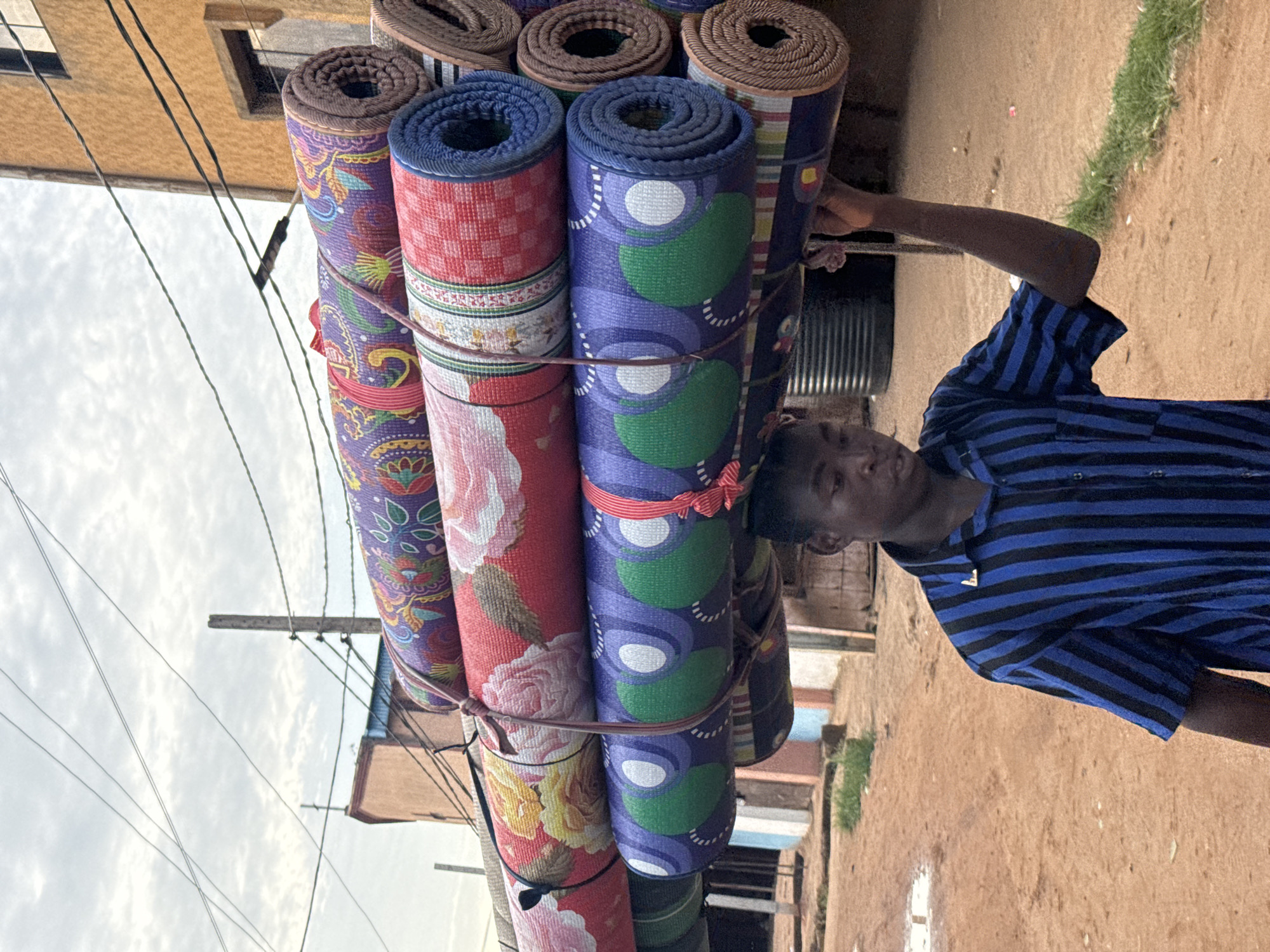 A young man in a blue and black striped shirt stands on a sandy, brown path in Onitsha, Nigeria. He carries a large stack of rolled mats on his head and back, secured by what appears to be string or rope. The mats have various colors and patterns, including purple with floral designs, red with floral patterns, and blue with green and white circular shapes. The surrounding environment shows buildings, utility poles with wires against a cloudy sky, and patches of green vegetation. The scene suggests a commercial activity, possibly the man selling or transporting the mats in an urban area.