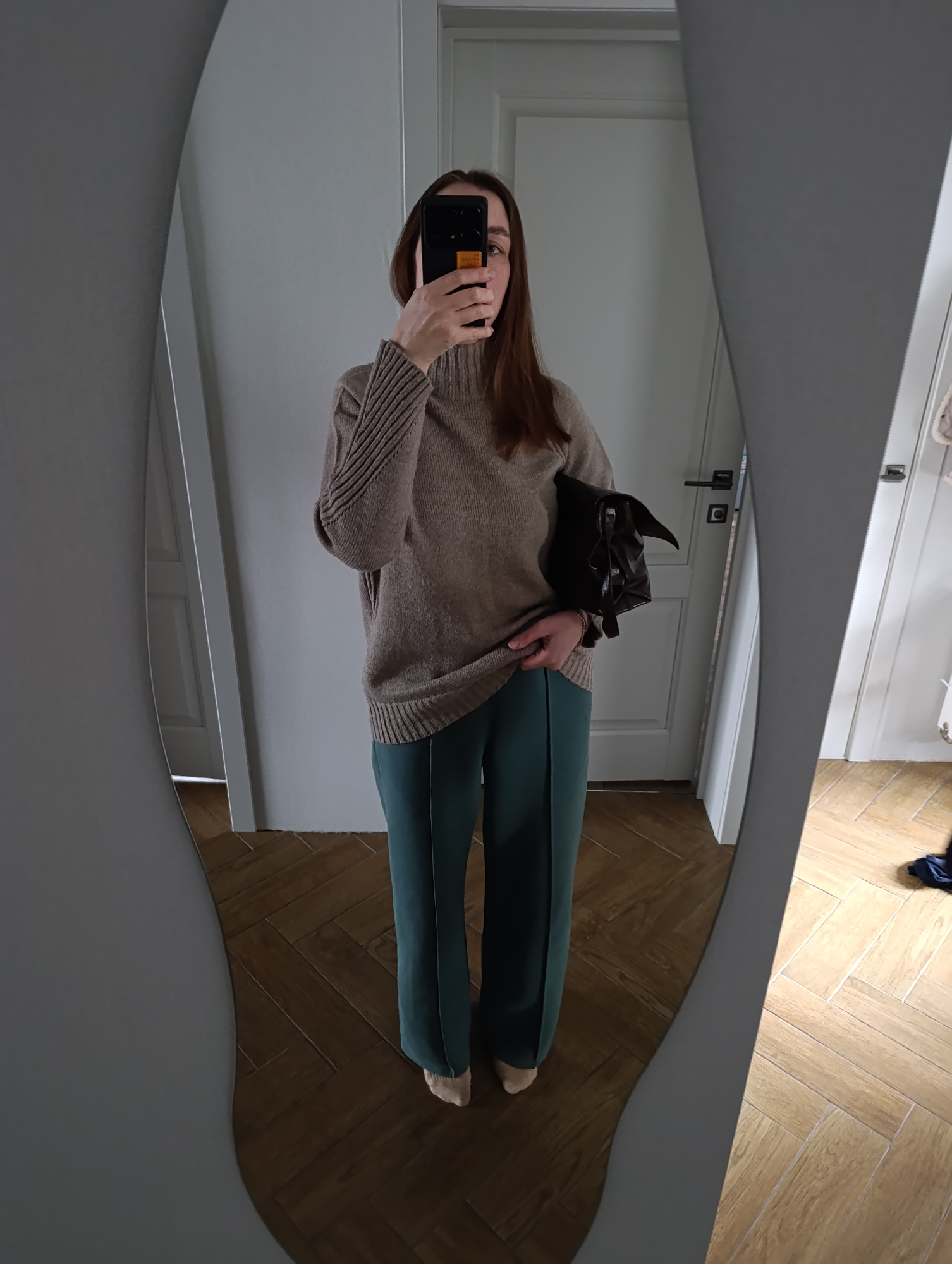 A mirror selfie captures a person indoors, presenting an outfit. The subject is a woman, whose face is obscured by the black smartphone she holds with both hands to take the picture. She is dressed in a cozy-looking, light brown or beige turtleneck sweater with ribbed detailing on the sleeves, paired with dark teal or deep green wide-leg trousers. Her feet are partially visible, wearing light-colored socks or slippers. A dark brown or black structured handbag is tucked under her left arm.
The reflection in the irregularly shaped mirror with a gray border reveals a clean, well-lit interior. Behind the woman, white doors and white walls are visible. The flooring is a light brown wooden parquet, laid in a herringbone pattern. On the back of the smartphone, there is a small rectangular orange or yellow sticker with some unreadable text. The overall scene suggests a residential setting, likely in Chaikovsky, Russia, during the daytime given the soft, bright ambient light.