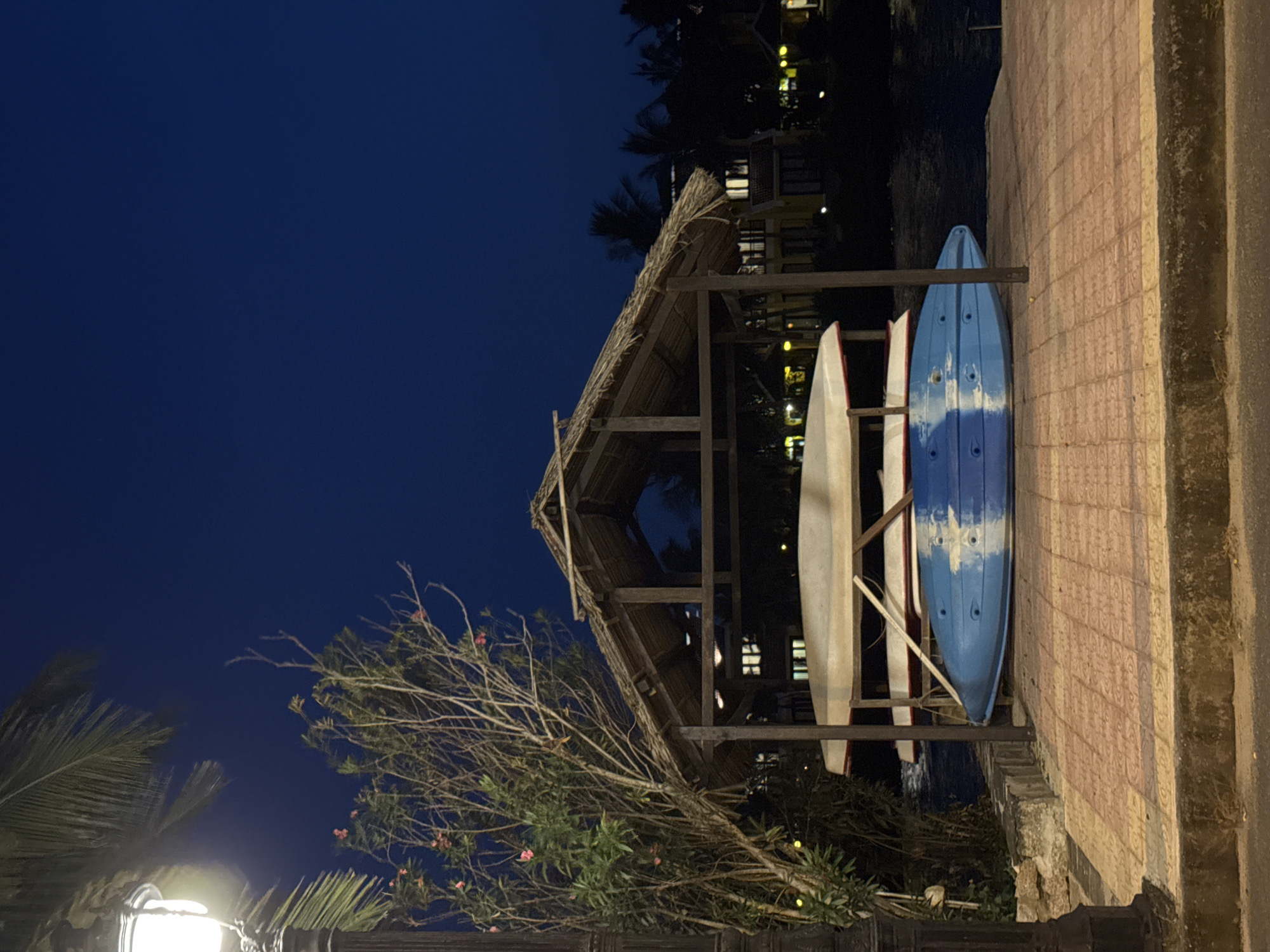 The image shows two kayaks, one blue and white, and one white, stored vertically under a thatched-roof shelter. The location appears to be coastal, possibly in Đà Nẵng, Vietnam, given the tropical vegetation and architecture. The scene is at night, with a deep blue sky. A tiled path or wall is on the right side of the image, running vertically. Trees and other vegetation are in the foreground. A lamppost provides light on the lower left. Other buildings are visible in the background.