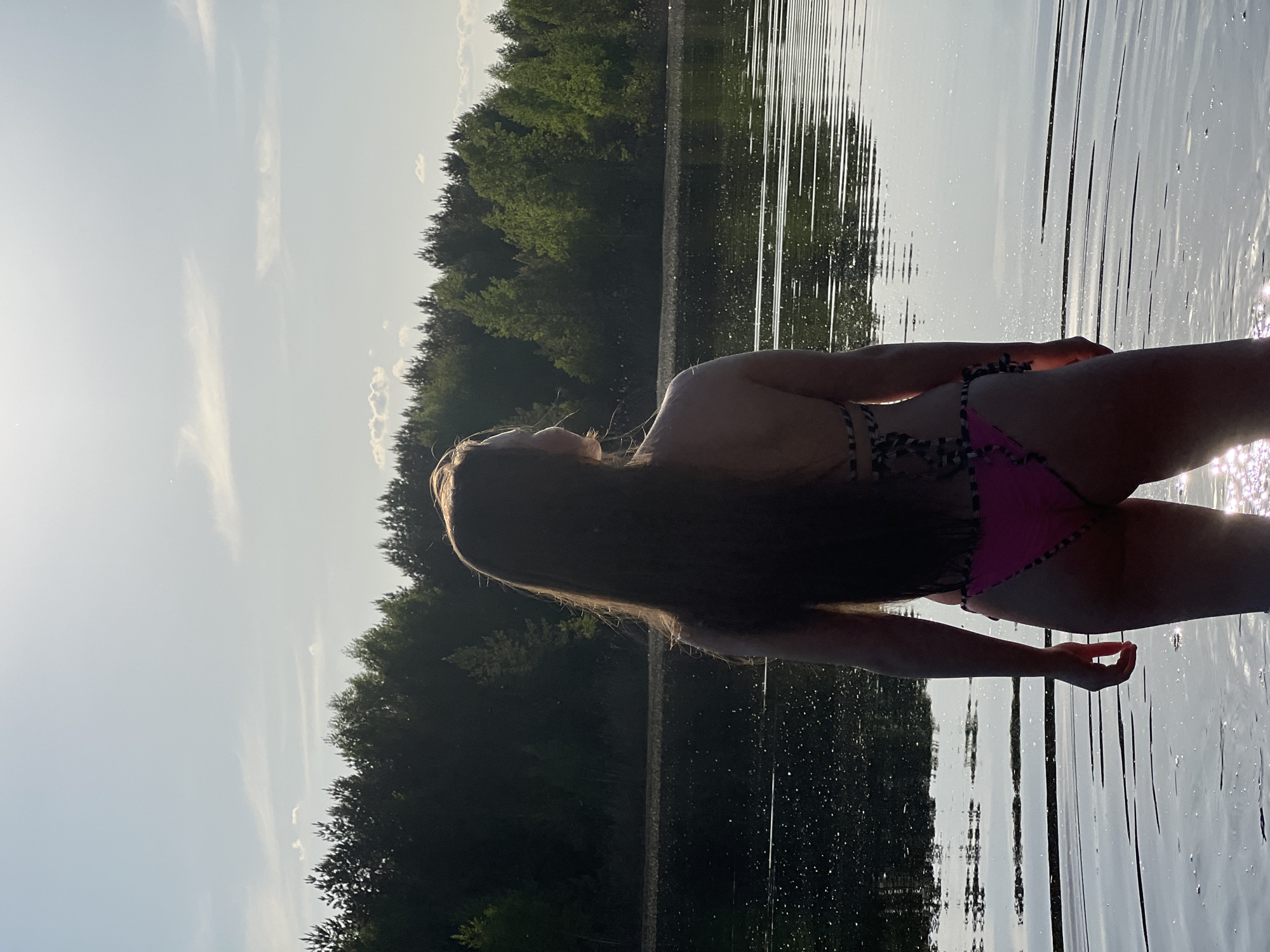 A woman is standing in a body of water, likely a lake, with a forest visible in the background. She is wearing a bikini with a pink bottom and a top with striped straps. The water is still and reflects the sky and trees. The overall scene suggests a summer day in a natural setting. The image does not provide the details necessary to confirm the location in the city of Novyi, Russia.