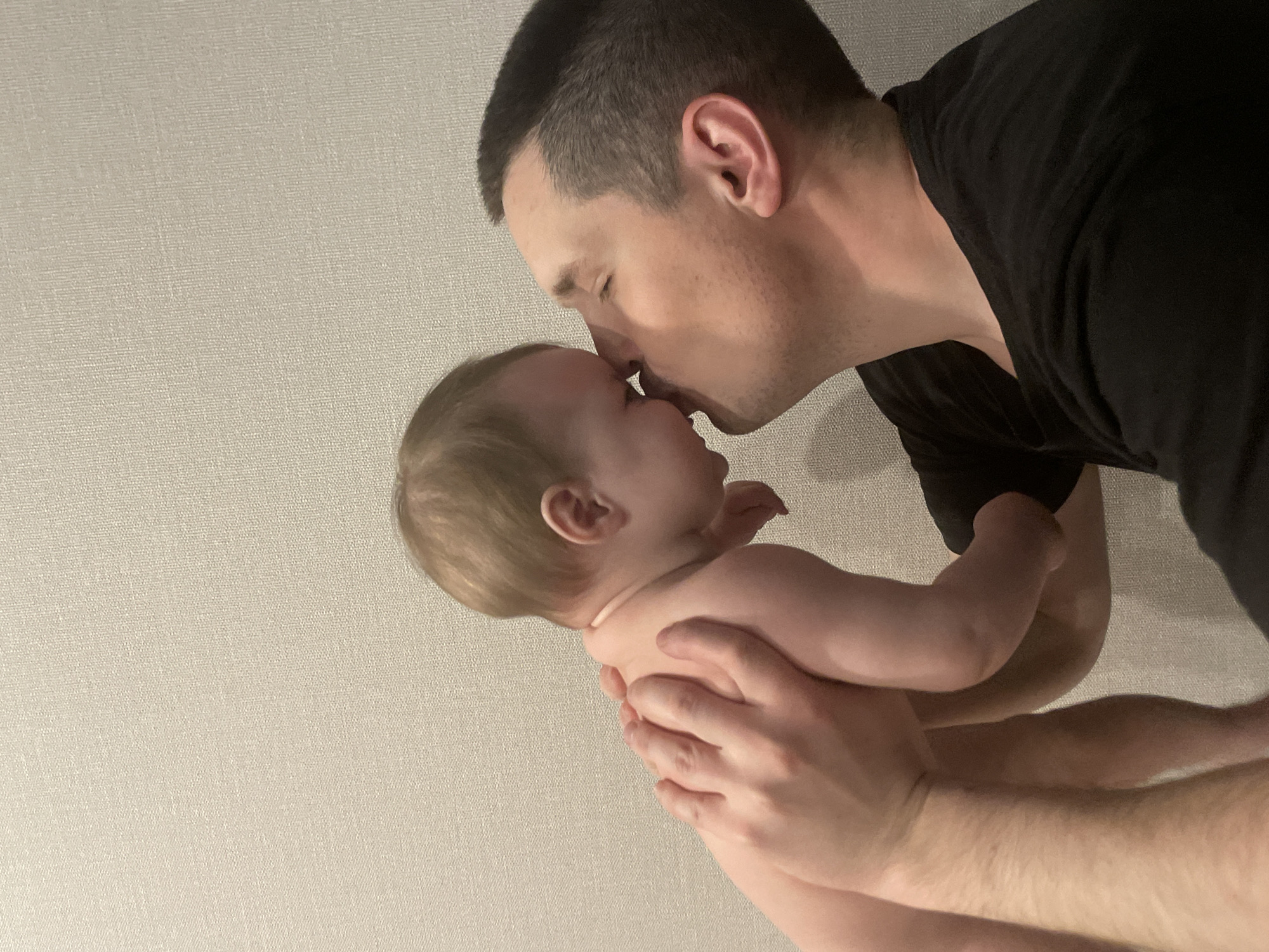 A man wearing a black shirt is kissing a baby on the nose. The background is a textured, light-colored wall. The location cannot be determined.