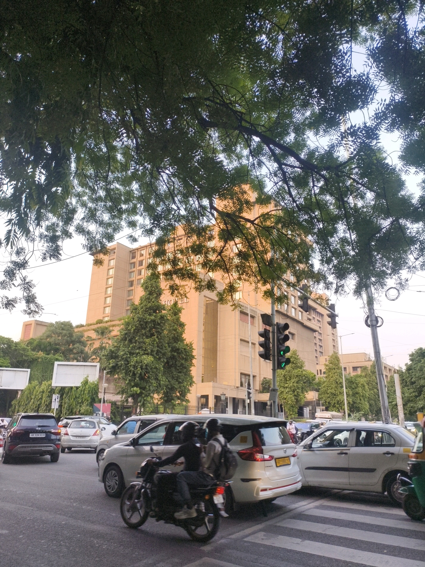The image shows a street scene in Delhi, India, with a large hotel building in the background. Several vehicles, including cars, a motorcycle, and an auto-rickshaw, are stopped at a traffic light. People are visible on the motorcycle and in one of the cars. Trees with lush green foliage overhang the street, partially obscuring the view of the sky. The traffic light is green, indicating that the vehicles may proceed. The hotel building appears to be a modern, multi-story structure. The overall impression is of a busy urban environment during daylight hours.