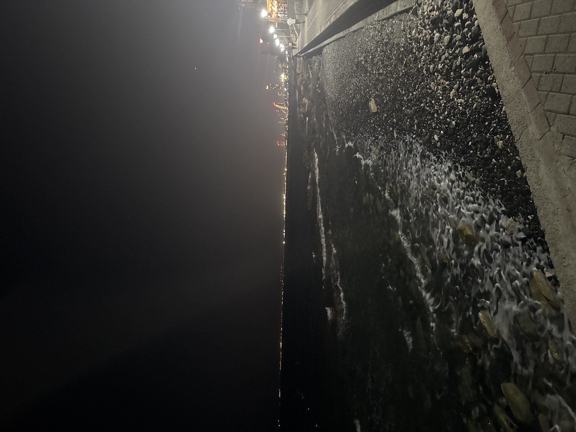 A nighttime scene captures a waterfront promenade in Agios Tychonas Community, Cyprus. The image looks out over a dark sea with small waves lapping against a rocky shore. In the distance, a long line of city lights stretches along the coastline, punctuated by illuminated skyscrapers. To the right, a paved promenade with patterned brickwork runs parallel to the sea. Streetlights illuminate the walkway, and a few people can be seen strolling. The sky above is dark, with a faint glow from the distant city and a few visible stars or distant lights.