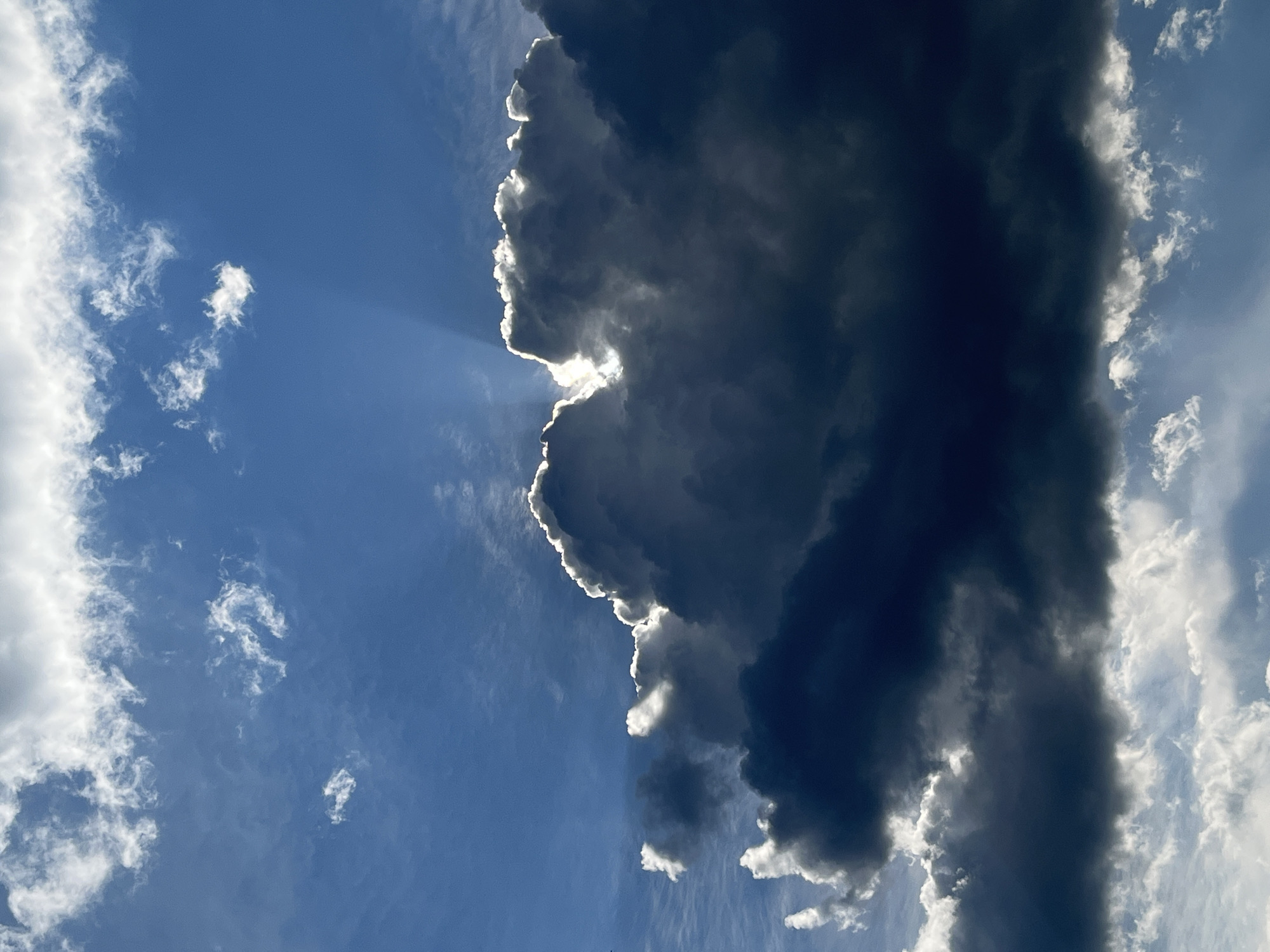 The image displays a dramatic sky filled with dark, heavy clouds, partially obscuring a bright blue sky. Rays of light emanate from behind the large central cloud formation, creating a halo effect and highlighting the edges of the clouds. Smaller, wispy white clouds are scattered throughout the upper and lower portions of the frame. The lighting suggests either sunrise or sunset, with the sun's rays piercing through the dense cloud cover. There are no people, structures, or distinct geographical features visible within this image, indicating it is solely focused on the atmospheric conditions. The image does not provide any information that would place it specifically in the city of Ореховка, Russia.