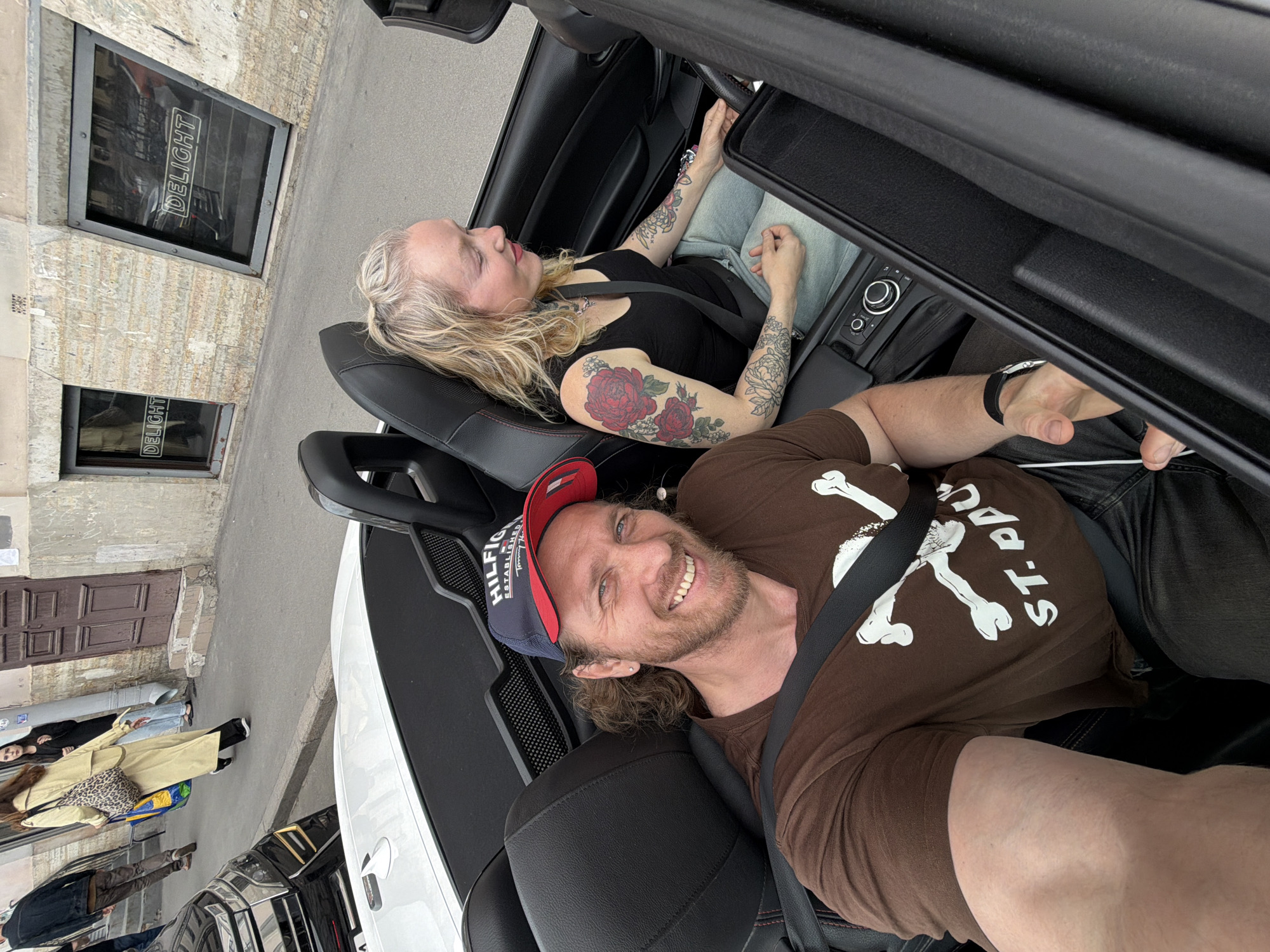 The image shows a man and a woman in a convertible car on a street in Saint Petersburg, Russia. The man is in the driver's seat and is smiling at the camera. He is wearing a brown t-shirt with a graphic print and a blue and red baseball cap. The woman is in the passenger seat and is looking to the right. She has blonde hair and is wearing a black tank top and light-colored jeans. She has tattoos on her arms. The car is black and appears to be a modern sports car. In the background, there is a building with windows that have the word "DELIGHT" written on them. The street appears to be cobblestone. There are other people and a black car visible in the background.