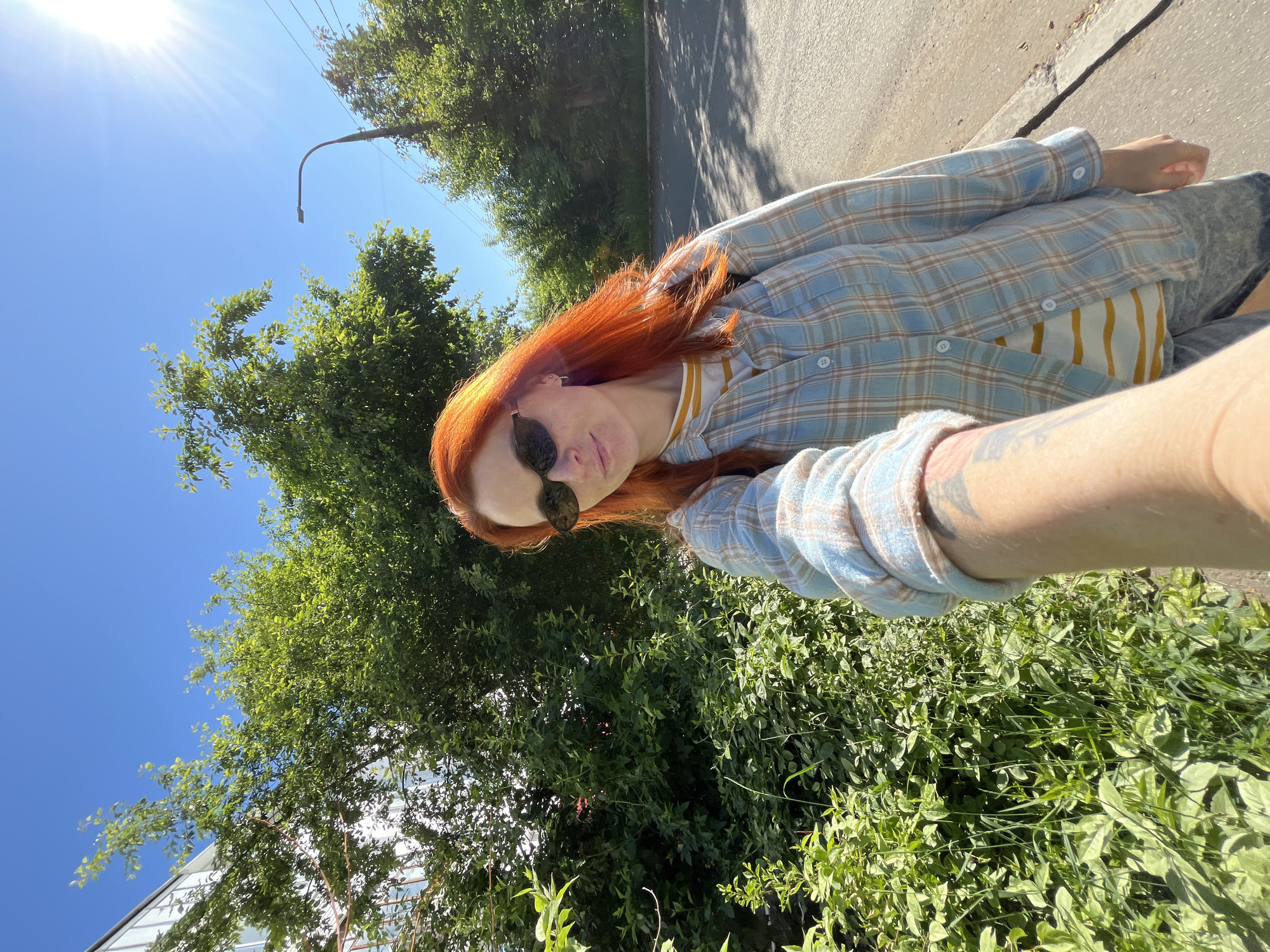 A woman with bright orange hair and sunglasses is taking a selfie outdoors on a sunny day. She is wearing a light blue and brown plaid button-up shirt over a yellow and white striped t-shirt, and denim shorts. Green trees and bushes are visible behind her, with a building on the left and a street with a sidewalk on the right. A lamppost and power lines are also present in the background. The scene appears to be in a residential area of Chaikovsky, Russia, given the architecture of the building and the overall urban greenery.