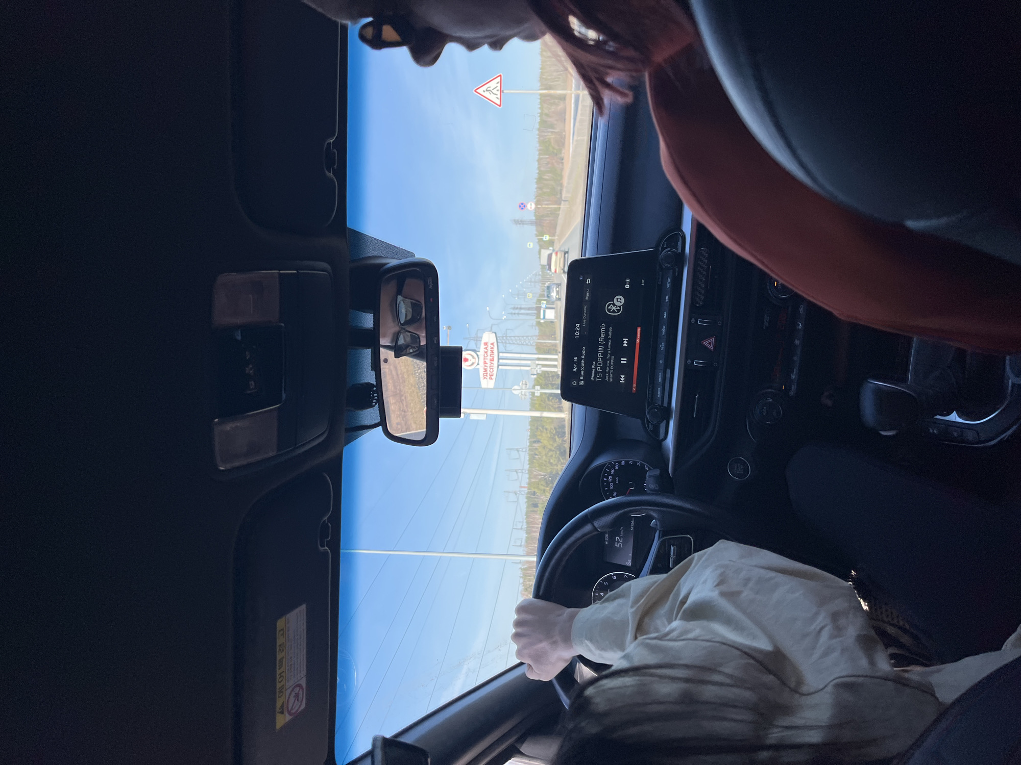 The view from inside a car, focused on the driver, with a glimpse of the road ahead. The vehicle is moving at 52 km/h. The infotainment system shows a song called "TS POPPIN (Remi)" by Jack Harlow, Tory Lanez, and DaBab. The dashboard displays a timestamp of 10:24 on April 16. A sign indicating "УДМУРТСКАЯ РЕСПУБЛИКА" is visible, suggesting this location is near or within the Udmurt Republic. Another sign with a pedestrian crossing symbol is also present. A person is in the passenger seat and the driver has their hands on the steering wheel.