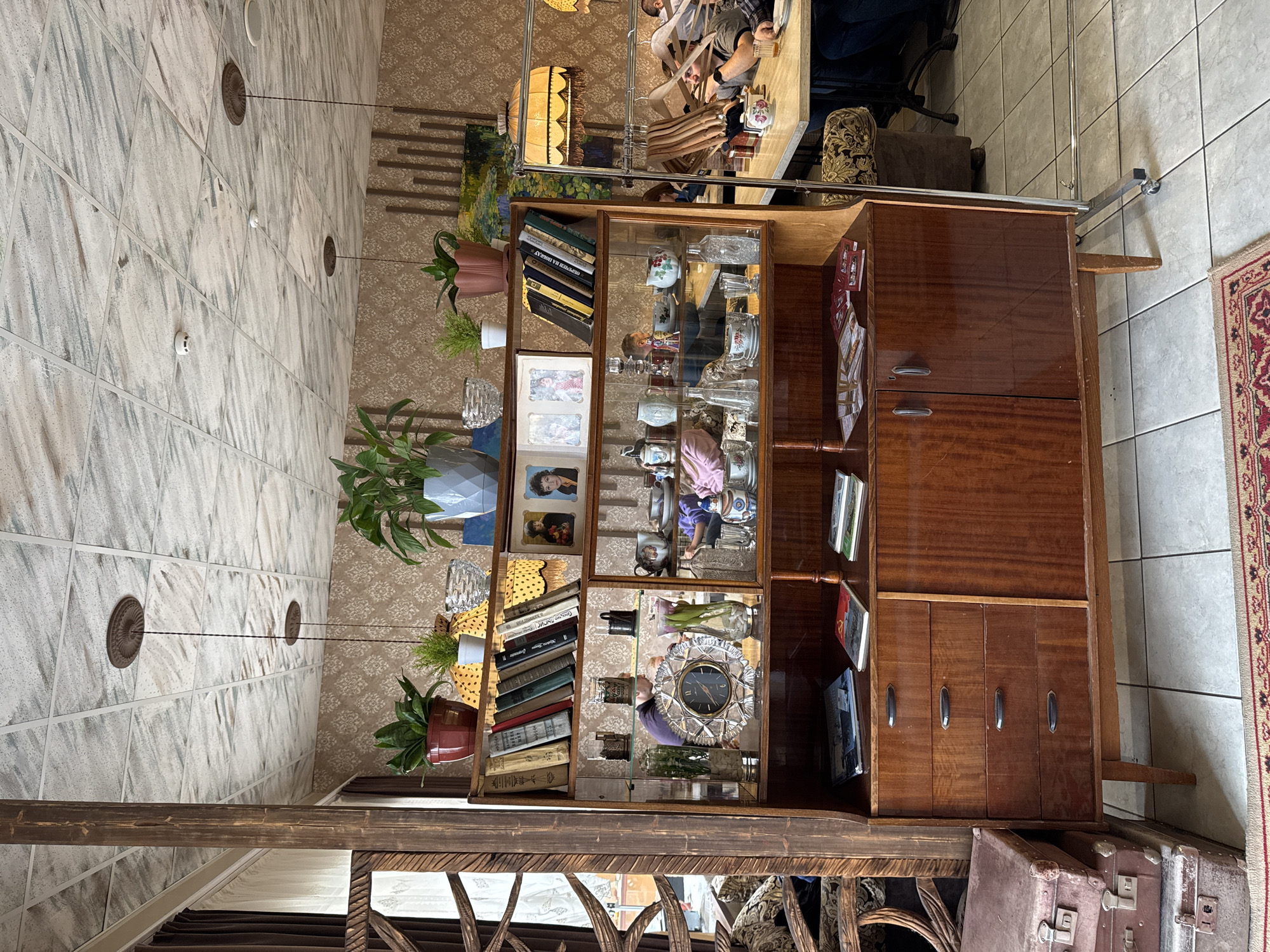 The image depicts the interior of a space in Kladovitsy, Russia. Dominating the scene is a large, dark wood shelving unit filled with an eclectic assortment of objects: books, photographs, decorative glassware, a clock, and houseplants. The unit has both open shelving and closed cabinets. A lamp is positioned above the shelving unit. A small table is visible, with people sitting at it. The flooring consists of light-colored tiles. The room has a patterned wallpaper and architectural details such as ceiling fixtures. A section of a rug with a red pattern is visible near the edge of the frame. Old suitcases are visible at the bottom of the image.