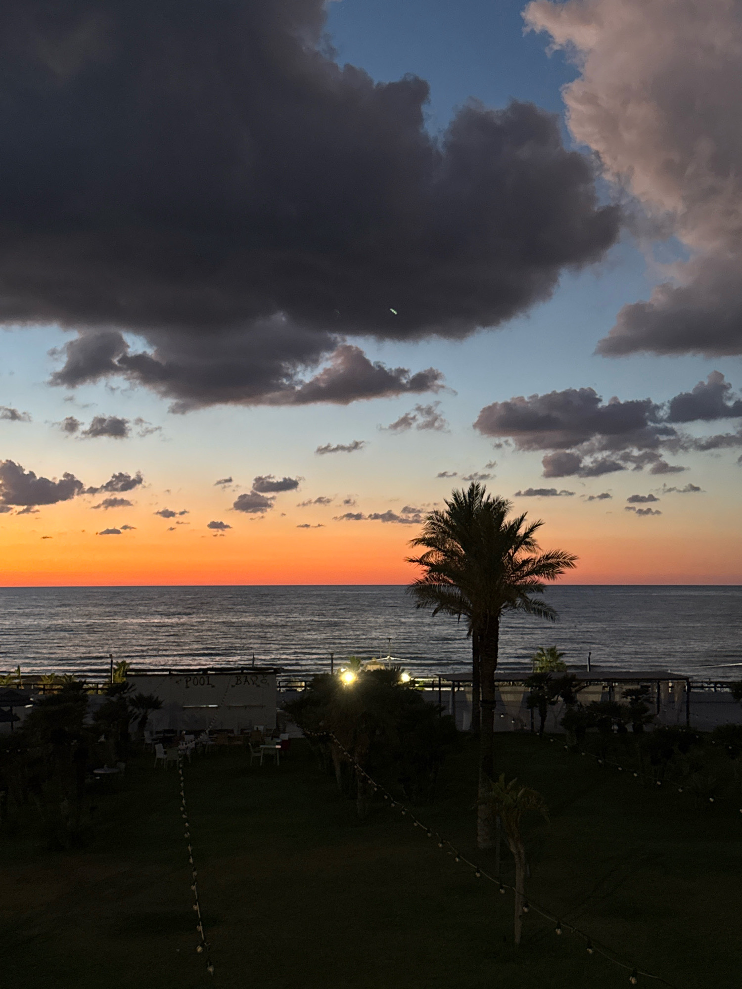A dusky sunset over the sea frames a scene on the coast of Isola delle Femmine, Italy. The sky transitions from a deep twilight blue at the top, speckled with dark clouds, to a vibrant orange and pink band near the horizon, where it meets the calm, dark ocean. A solitary palm tree stands silhouetted against the colorful sky in the mid-ground, its fronds catching the fading light.
In the foreground, a manicured lawn is sparsely populated with small, decorative trees. Further back, near the water's edge, is a structure with "POOL BAR" visible on its facade. White tables and chairs are arranged in front of it, suggesting an outdoor dining or lounge area. Strings of small lights are draped across the scene, some visible on poles and others winding through the grass, casting a dim glow in the encroaching darkness. The overall impression is of a peaceful, post-sunset ambiance at a coastal establishment.