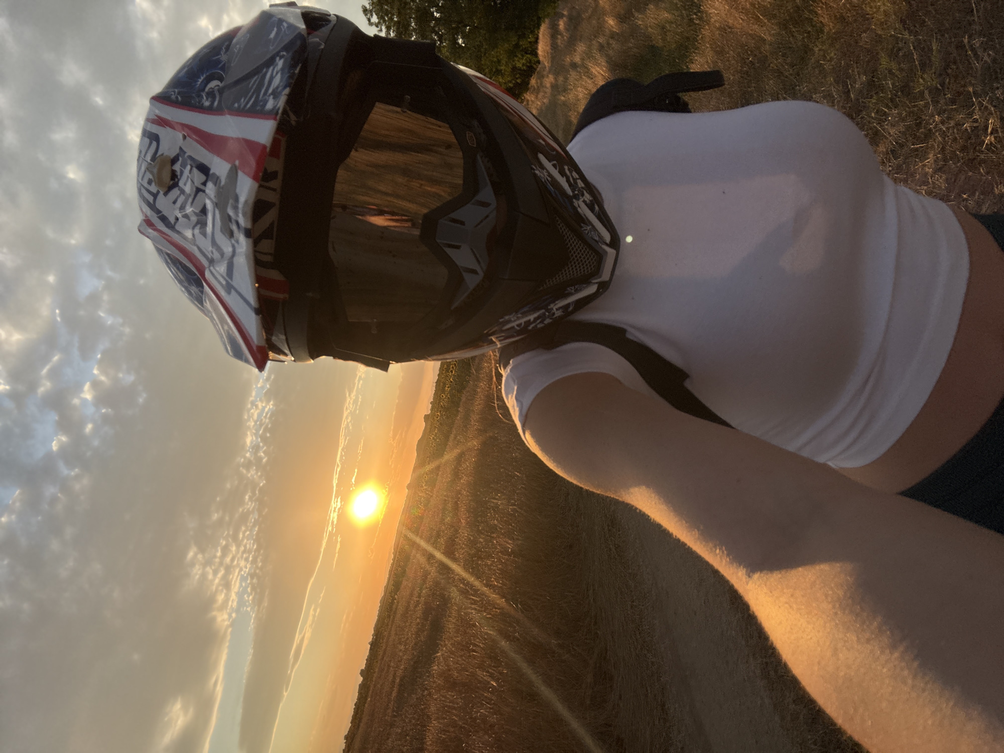 A first-person perspective selfie is taken in a rural field at sunset. The subject is wearing a white t-shirt and a motocross helmet with goggles. The sun is setting on the left side of the image, casting a warm glow over a field of dry grass and some sunflowers. The sky is filled with clouds illuminated by the sunset. The location is identified as the city of Stroevo, Bulgaria.