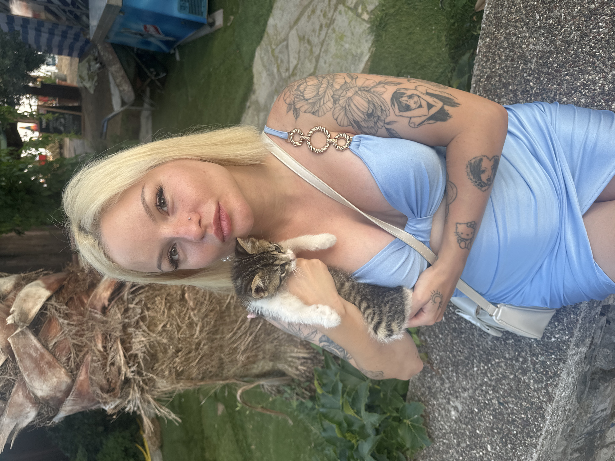 A woman with blonde hair and tattoos is holding a small tabby kitten. She is wearing a light blue dress with gold ring details on the strap. A light-colored purse is visible. The woman is holding the kitten close to her chest. The scene appears to be outdoors, with greenery and a textured stone surface in the background. The image suggests a casual, possibly warm-weather setting.