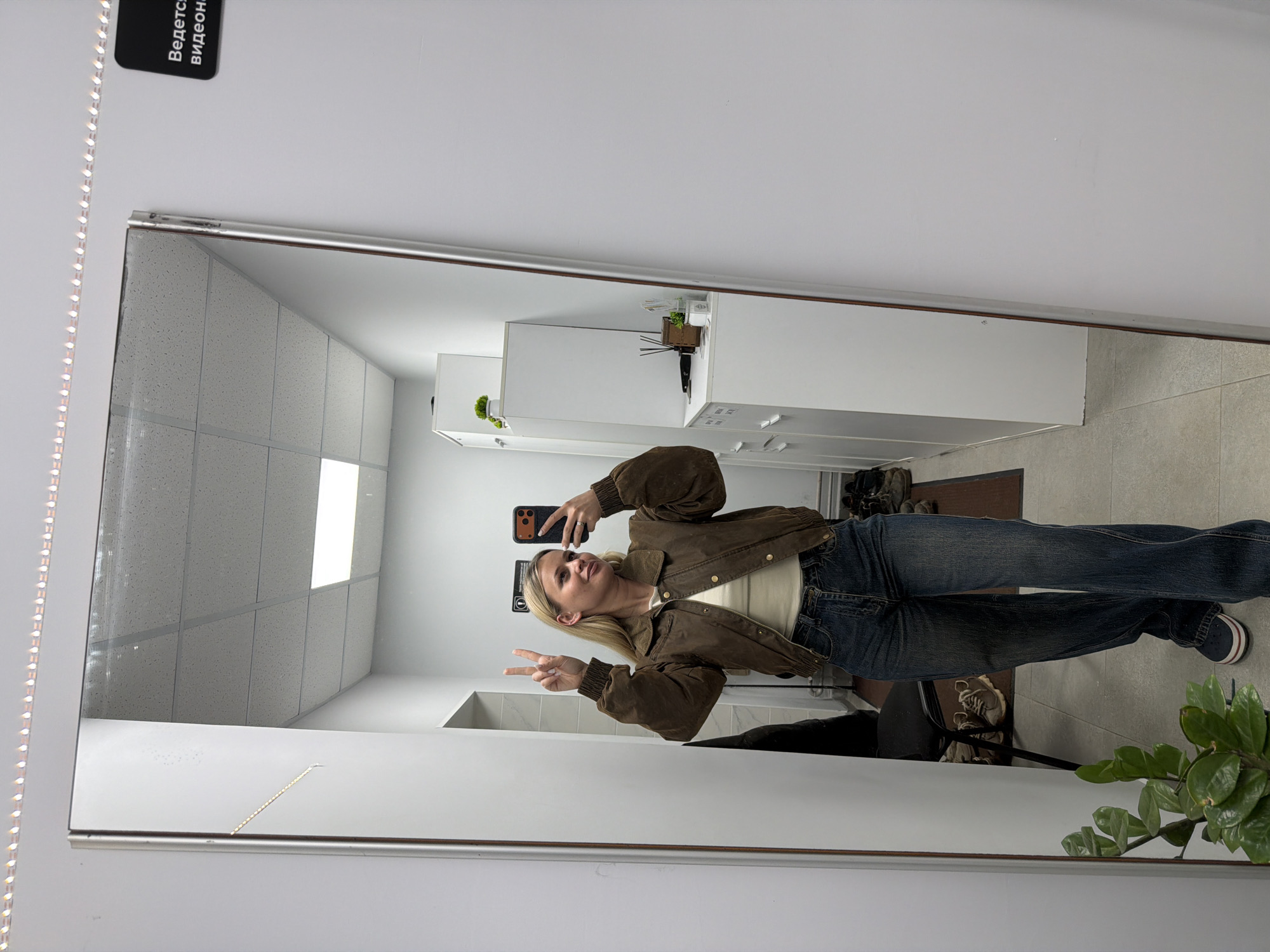 The image is a mirror selfie of a blonde woman in blue jeans and a brown jacket, holding a phone and making a peace sign. The background shows an interior space with white walls, tiled ceiling, white cabinets, and a plant in the corner. It is difficult to determine if the image was taken in Sarapul, Russia, without additional context or information.