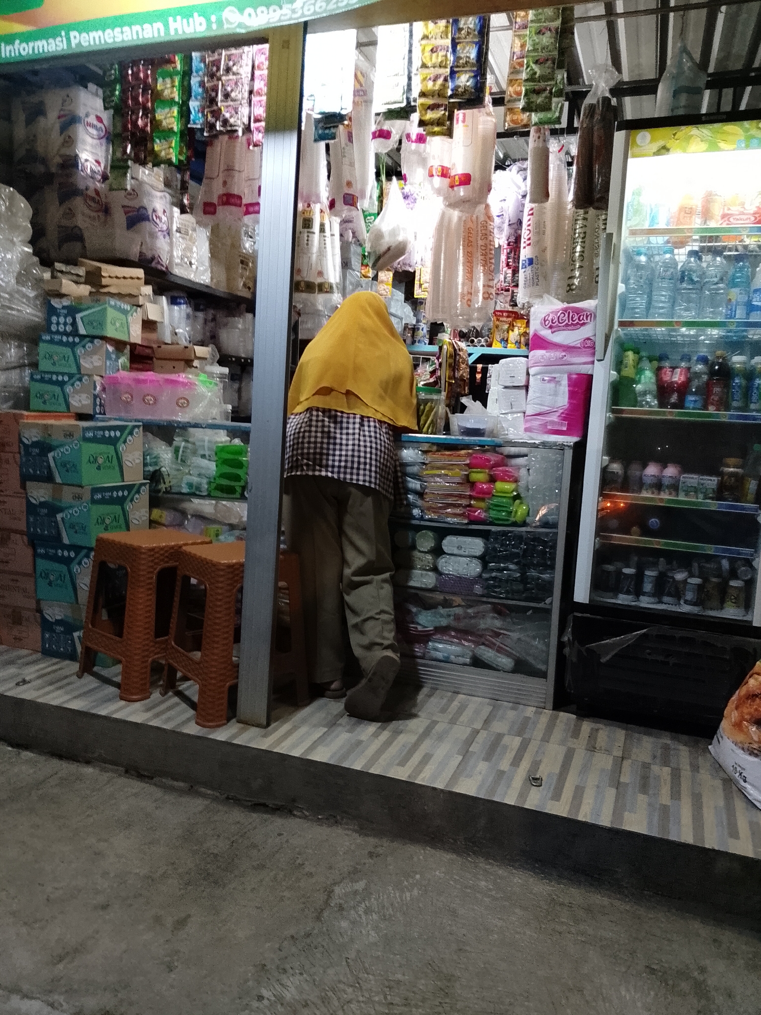 The image depicts a storefront in Pasuruan, Indonesia, displaying various goods for sale. A person wearing a yellow hijab and a checkered shirt is partially visible, facing away from the camera and interacting with the merchandise. The storefront is stocked with an assortment of items, including plastic cups, packaged food items, and beverages. A refrigerator stocked with drinks is visible on the right side of the frame. The floor is tiled, and concrete surrounds the entrance. The overall scene suggests a small retail establishment in an Indonesian city.
