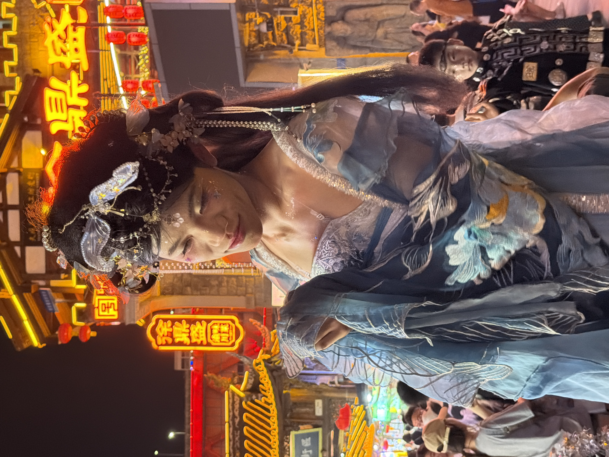 The image captures a nighttime scene in the Yuzhong District of China, illuminated by vibrant neon signs. In the foreground, a person dressed in an elaborate, flowing blue and white costume with floral embroidery and intricate hair ornaments is the primary subject. Their face, with glitter makeup, is tilted downwards, suggesting contemplation or engagement with something below. The costume has a traditional, possibly theatrical, aesthetic.
In the background, other people are visible, some appearing to be spectators or participants in an event. One person in the mid-background wears a black outfit with a distinctive pattern, suggesting they might also be in costume. The setting appears to be a bustling street or plaza, with shops and establishments adorned with bright, colorful lights. Red lanterns are also visible, adding to the festive atmosphere.
The visible text on the neon signs includes Chinese characters. One prominent sign in yellow characters reads "悠遊居" (Yōuyóu jū), which translates to "Leisurely Residence" or "Relaxing Place." Another sign in yellow and red characters reads "渝中" (Yúzhōng), which is the name of the district. The overall mood is lively and energetic, characteristic of a night market or cultural event. The time of day is clearly night, with the dark sky contrasting sharply with the artificial illumination.