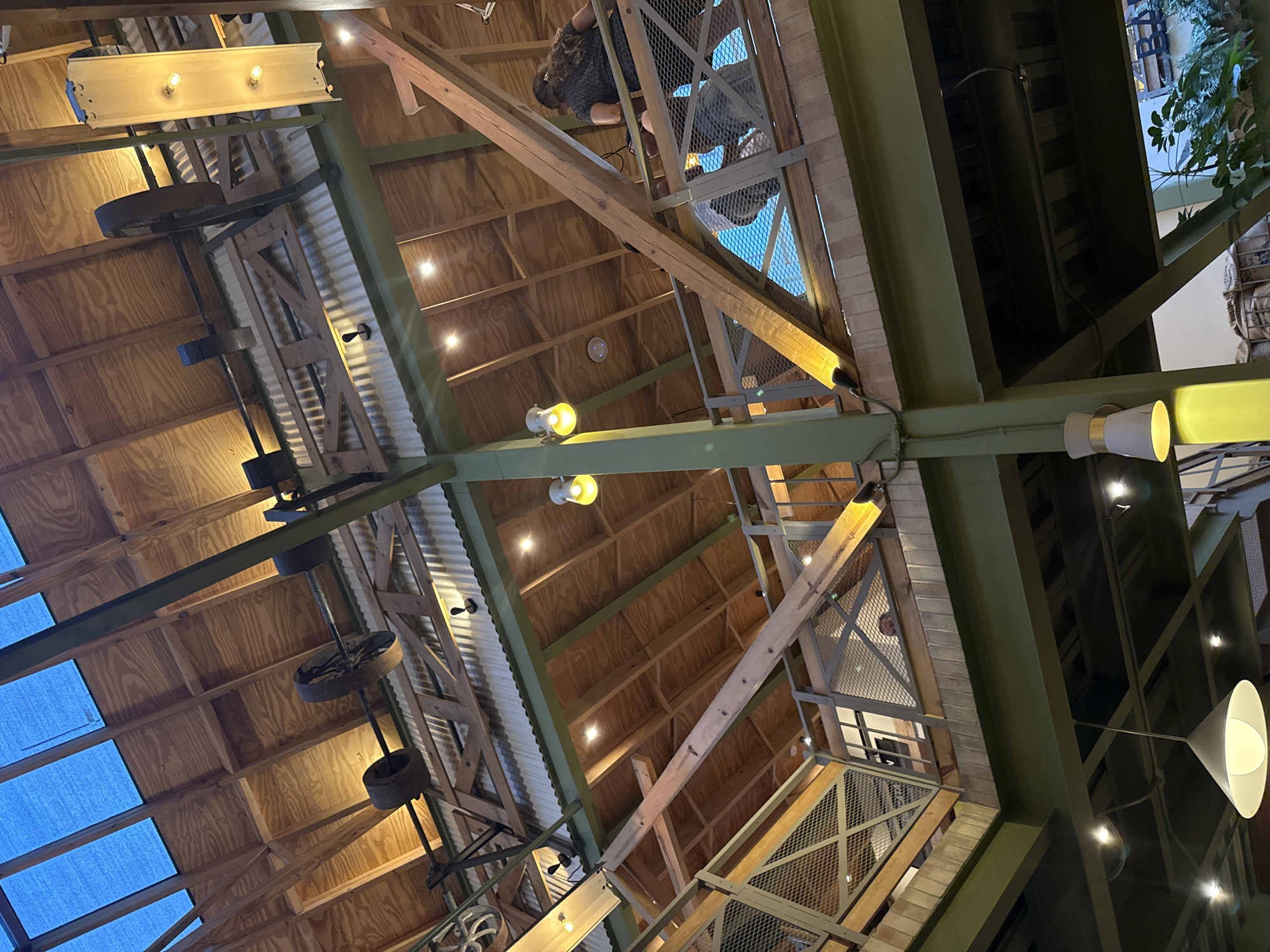 The image depicts a low-angle view of a building's interior, possibly a commercial space or restaurant, in San Cristóbal, Mexico. The architectural structure features an exposed wooden beam ceiling with sections of corrugated metal roofing. Industrial-style lighting fixtures are present, including exposed bulb lamps and hanging cone lights. A decorative arrangement incorporating repurposed industrial items, such as metal wheels or gears, is suspended from the ceiling. The building structure also includes green-painted metal beams and a multi-level design with metal railings and grating, suggesting a mezzanine or platform area. An individual or individuals can be seen on one of the upper levels. There are also potted plants and other decorative elements in the space.