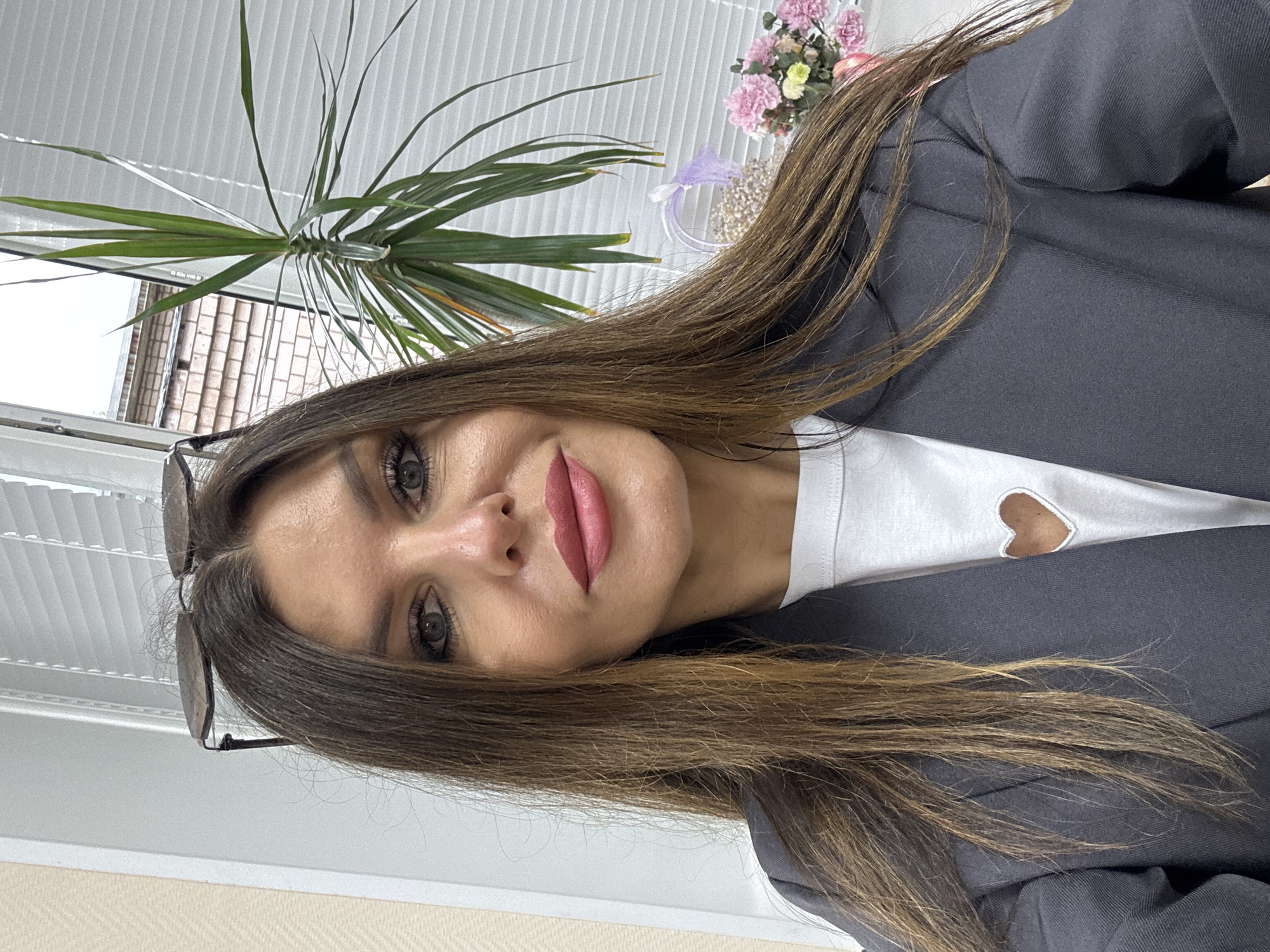 A head-and-shoulders portrait features a woman with long, straight brown hair, dark eyes, and pink lipstick, looking directly at the camera with a slight smile. She wears a grey blazer or jacket over a white t-shirt that has a heart-shaped cutout or design near the neckline. Sunglasses are resting on top of her head.
The setting is indoors, with a window covered by white venetian blinds visible behind her. Through the window, parts of a brick building can be seen, indicating an outdoor urban or suburban environment. An indoor plant with long, spiky green leaves is positioned in the background near the window. To the right of her head, a small decorative arrangement of artificial flowers, primarily pink and white with hints of purple, is visible. The lighting suggests it is daytime. The woman appears calm and composed. The location context for the image is Chaikovsky, Russia. A blurry white rectangular object visible near the window in the background may have indistinct text, possibly "MIMMO".