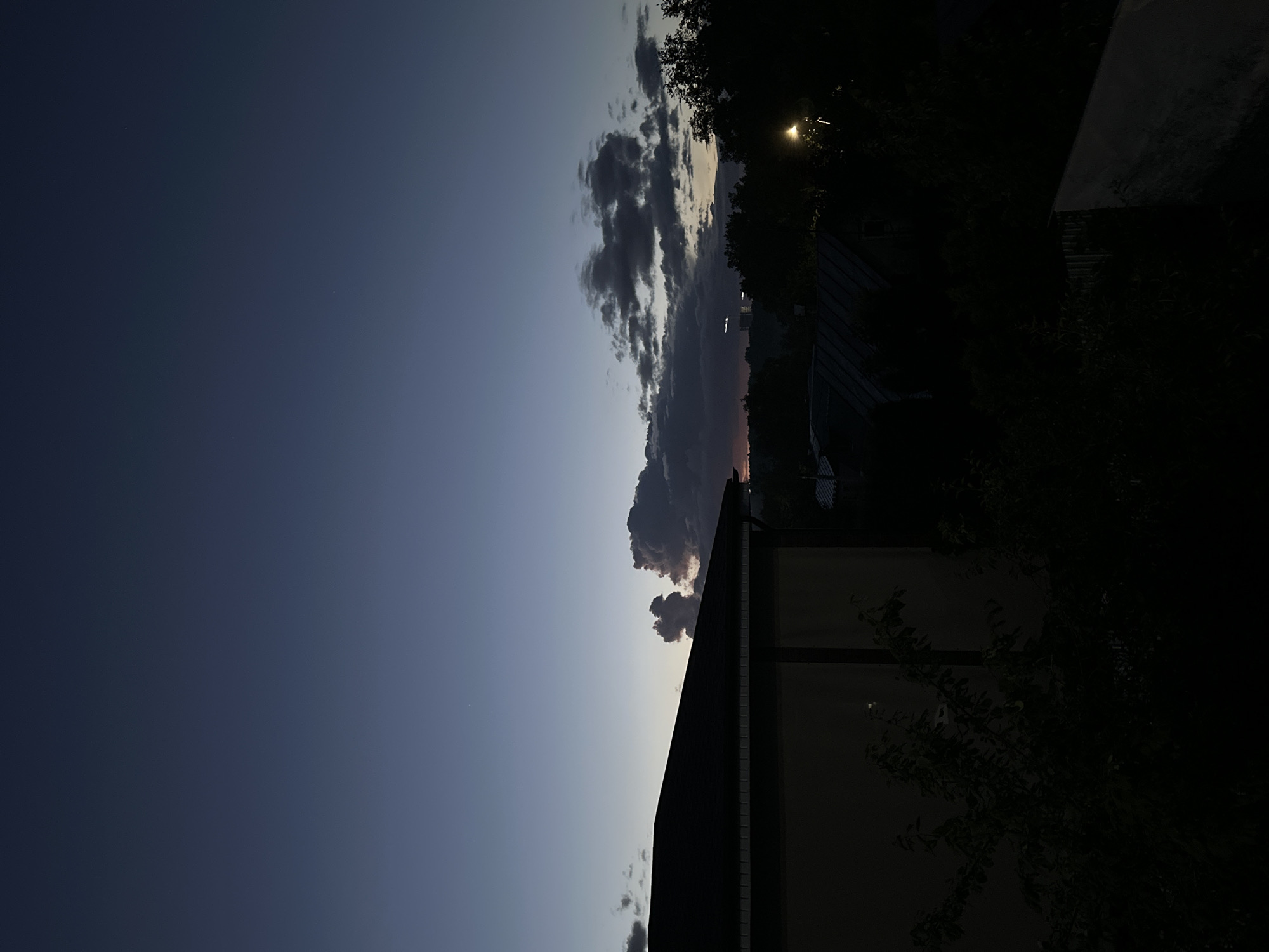 The image depicts a twilight scene over a residential area. In the foreground, the dark roof and upper wall of a building are visible on the left. The middle ground is filled with silhouetted trees and rooftops, with a street lamp providing a warm light source on the right. In the background, a faint glow on the horizon suggests the last remnants of sunset, with some distant buildings barely discernible. The sky is a deep, fading blue, transitioning to a darker hue at the top, with scattered dark clouds. The overall atmosphere is calm and serene, characteristic of dusk in a suburban or semi-urban environment. The location is the city of Nizhny Novgorod, Russia.