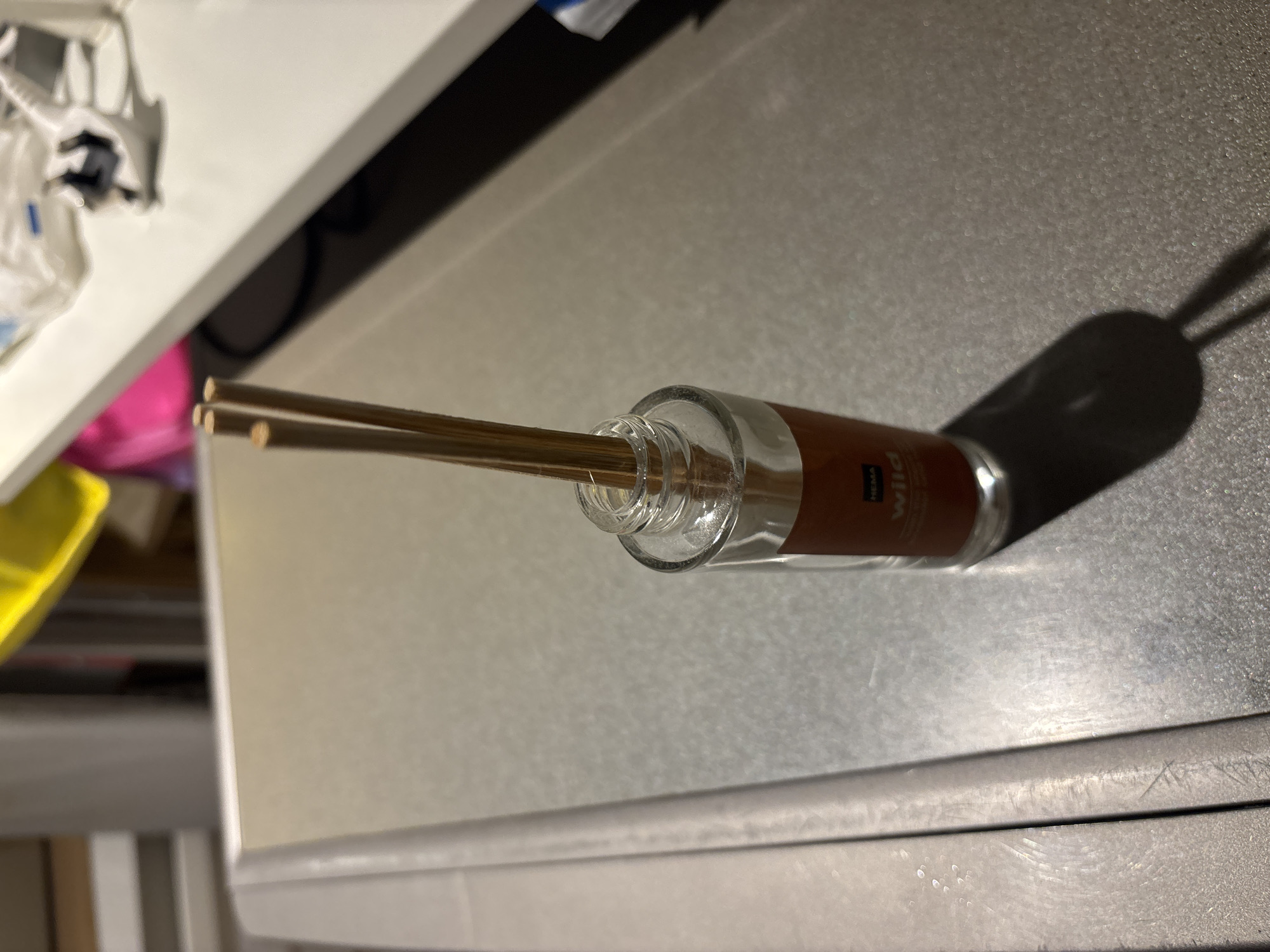 The image displays a close-up, high-angle shot of a small glass diffuser with wooden reeds inserted into it. The diffuser has an orange label with text that appears to read "HEMA" and "Wild". The diffuser is positioned on a textured, metallic surface that reflects light. In the background, a white shelf is visible with some blurred items, including what looks like a yellow object, a pink object, and some white plastic packaging with a black and white object. The overall lighting suggests an indoor environment, possibly a home or retail setting. There is no discernible connection to Dubai, United Arab Emirates, in the image content.