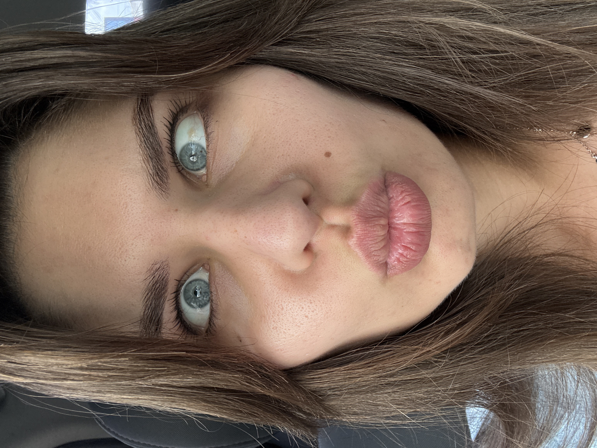 A close-up, head-on, medium shot captures a young woman's face from the mid-forehead to just below her chin. She has long, dark brown hair that frames her face and falls over her shoulders. Her eyes are wide open and looking in opposite directions, a common expression for a "cross-eyed" or "googly-eyed" look. Her lips are pursed into a puckered expression, as if for a kiss. A small, dark mole is visible on her right cheek, just below her eye. Her skin appears to be fair with visible pores. The background is out of focus, suggesting she is indoors, possibly in a car given the dark seat visible behind her. The lighting is bright, illuminating her face evenly.