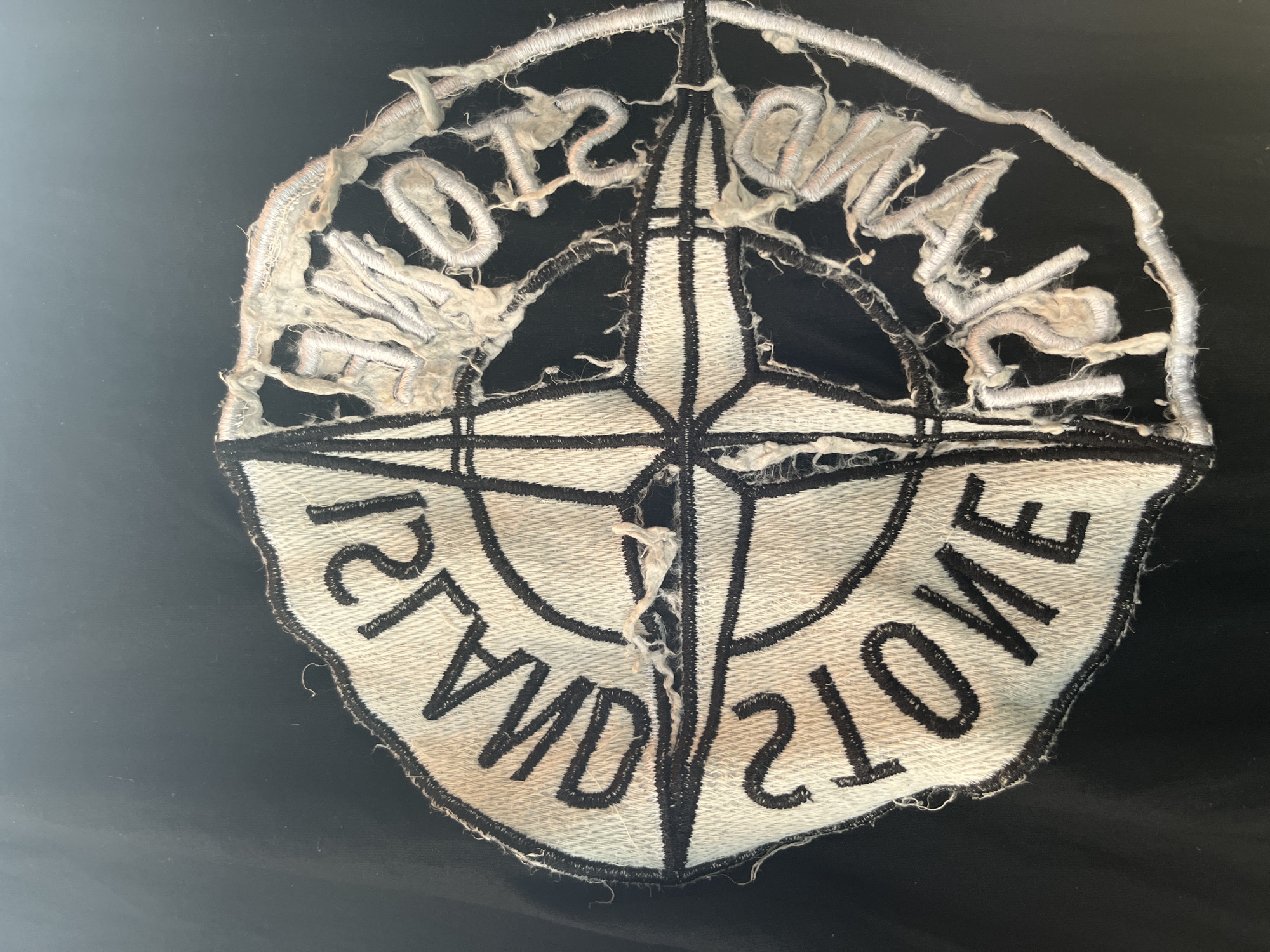 A worn and torn black and white circular patch is centered on a black fabric background. The patch features a compass rose design with the words "STONE ISLAND" embroidered in black text around the outer edge. The right half of the patch is significantly frayed and damaged, with loose threads and some of the embroidery missing. The left half of the patch is in better condition, with intact stitching and clear lettering. The image is a close-up shot, and no other elements of the environment are visible. The location in Central Jakarta, Indonesia, is not discernible from the provided image.