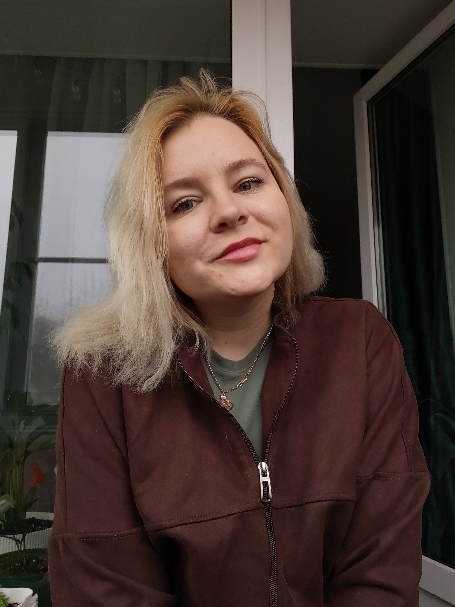 The image shows a woman with blonde hair, wearing a brown jacket over a green shirt and a necklace. She is indoors, likely in a room with a window overlooking a rainy day, indicated by the window's reflection. The location is Izhevsk, Russia, although specific buildings or landmarks are not visible in this close-up view. The details of the surrounding environment are limited, with a potted plant visible in the lower left corner. The woman is looking at the camera and slightly smiling.