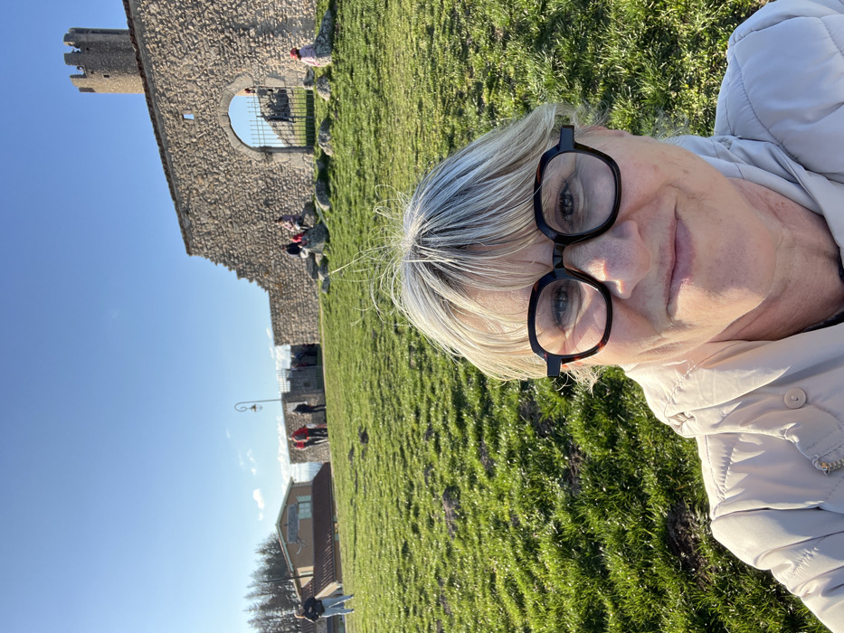 The image presents a close-up selfie of an adult female in the foreground, positioned outdoors under a clear blue sky in Chambles, France. The woman has short, light blonde hair, styled with bangs and visible flyaways at the crown. She wears large, dark tortoiseshell-patterned eyeglasses and a light beige quilted jacket, displaying a closed-mouth smile.
Behind the woman, a vibrant green grassy slope extends into the midground. To the left, a light-colored building with a dark roof and an unreadable sign is partially visible, with coniferous trees in the background. A solitary individual stands on the grassy slope near this building. A black, curved streetlamp stands to the left of a prominent stone wall.
The central background features a substantial stone wall, identifiable as part of the Château de Chambles, which includes an arched gateway. A tall, cylindrical stone tower with crenellated battlements rises behind the wall on the far right. Multiple individuals are present in the background: two people are seated on rocks near the wall to the left of the arch, another person wearing a pink/red hat is seated on the right, and several other figures are standing or moving near the wall and through the archway. The scene is brightly illuminated by direct sunlight, casting distinct shadows on the grass.