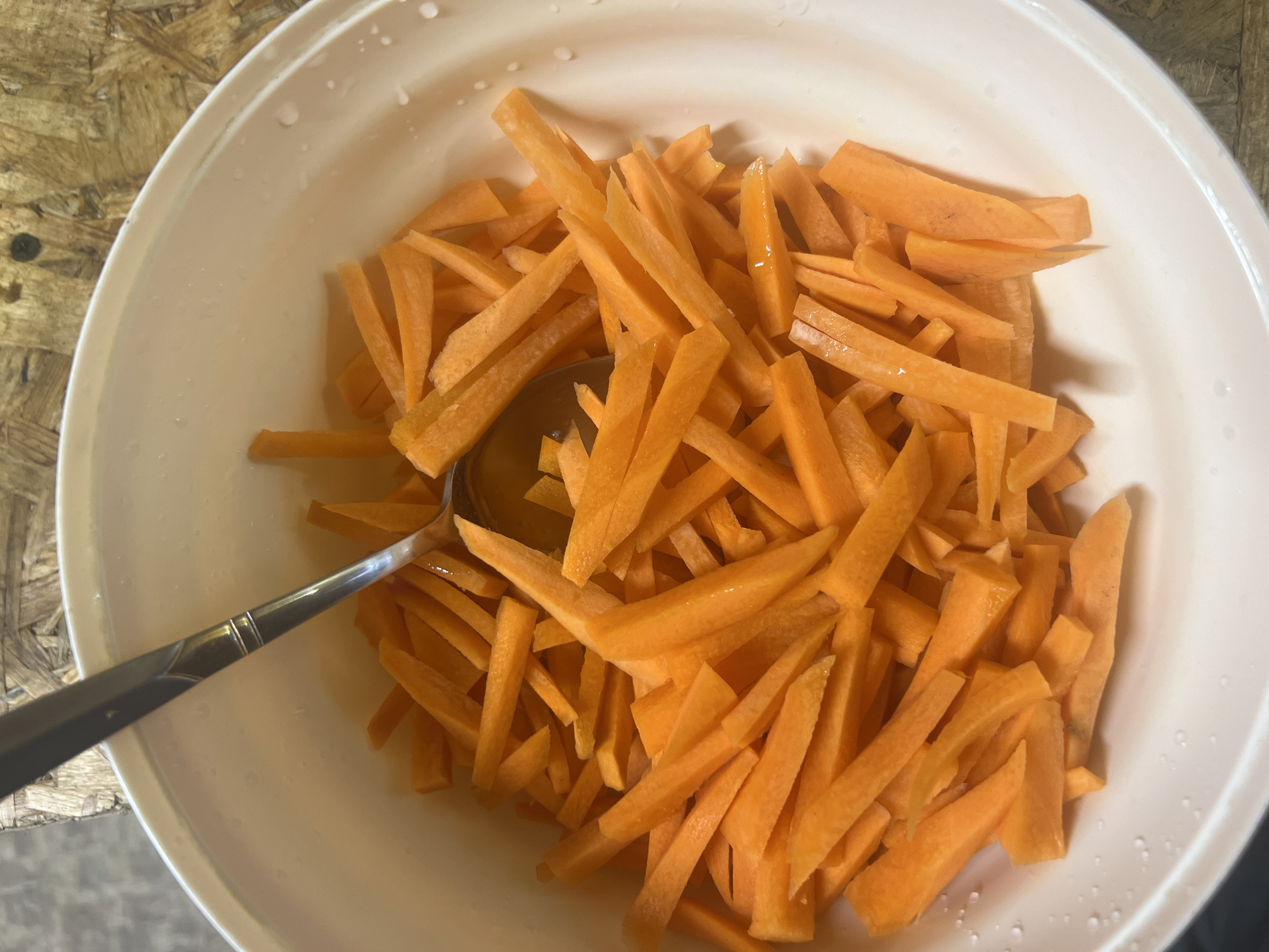 A close-up overhead shot shows julienned carrots in a white ceramic bowl. A silver spoon rests within the bowl, partially submerged in the carrots, with its handle angled upwards and to the left. The bowl is set on a textured, light brown surface that appears to be wood. Small droplets of water are visible on the inside rim and exterior of the bowl. There are no people or animals present. The scene is indoors, with the lighting suggesting it is daytime. The specific location in Yurlovo, Russia is not discernable from the image.