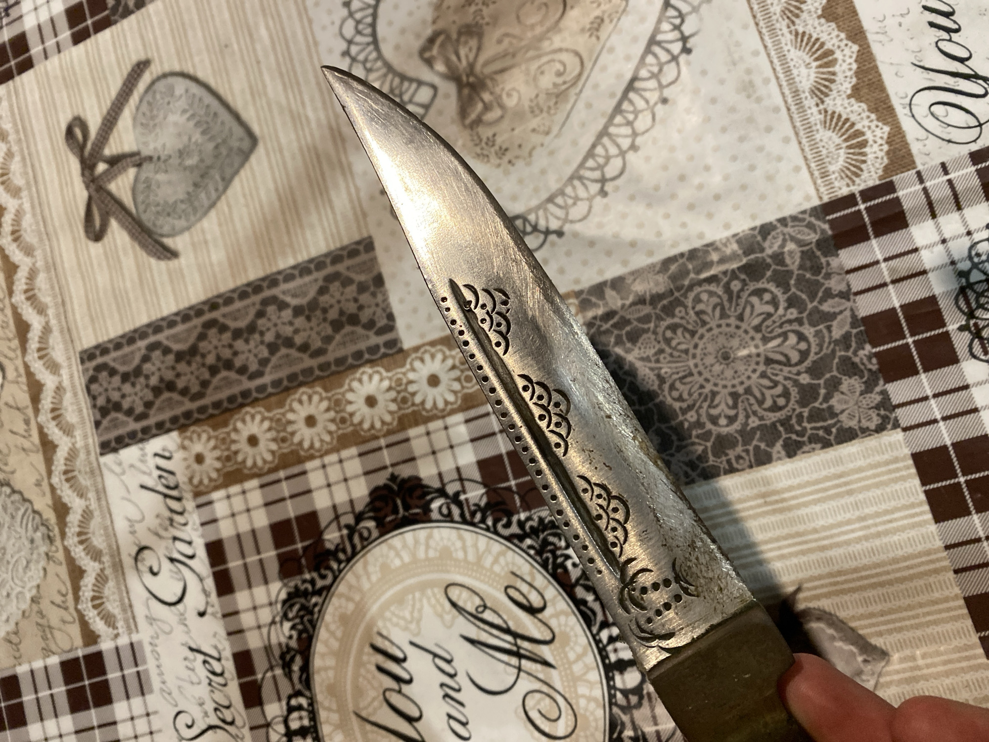 A close-up image shows a knife blade, its edge oriented vertically, held by a hand. The blade has a decorative design of evenly spaced circular perforations and stylized floral cutouts along one edge. The blade appears to be made of metal with a brushed finish. The background is a patchwork-patterned fabric with sections featuring hearts, floral designs, lace, plaid, and script. The overall composition suggests a domestic setting. There is no information to suggest that the image was taken in the city of Konstantinovo, Bulgaria.