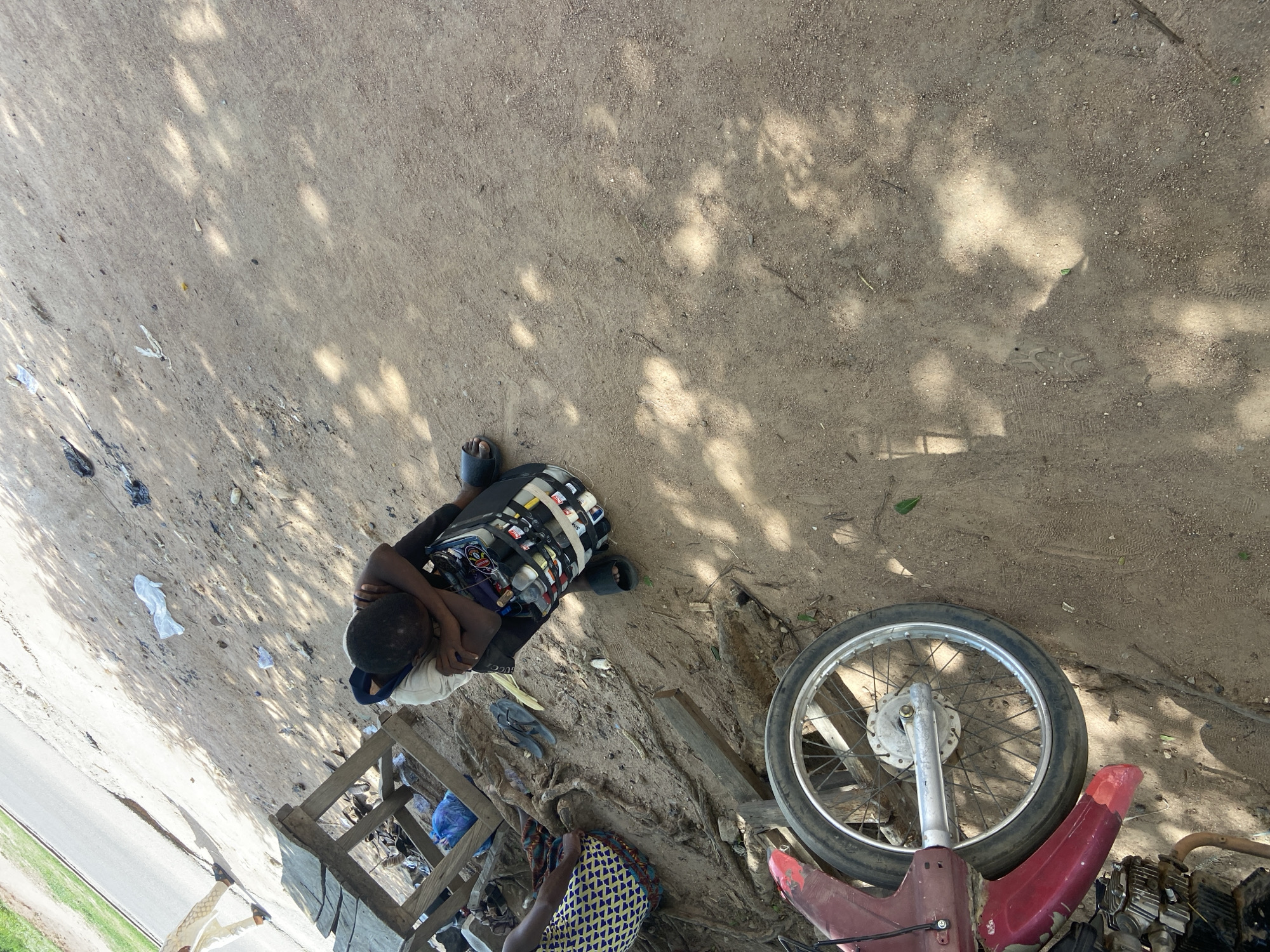 The image depicts a scene in Jalingo, Nigeria, under the dappled shade of trees. A person, lying down, is the central subject. They are wearing a dark t-shirt and a blue cap, with their head resting on their arms. The person appears to be asleep or resting. Scattered around are various objects, including plastic bags, debris, and what looks like discarded items.
In the foreground, parts of a red motorcycle are visible, including a wheel with spokes. A wooden structure, possibly a makeshift bench or stand, is also present. Another person is partially visible in the lower right corner, wearing a patterned fabric. The ground is dusty and appears to be a dirt or gravel surface, common in outdoor settings in many parts of Nigeria.
The lighting suggests it is daytime, with sunlight filtering through the leaves overhead, creating patterns of light and shadow on the ground. The overall impression is one of a casual outdoor setting, perhaps a roadside or a less-developed area, where people might rest or a motorcycle is being repaired or stored. There is no visible text in the image.